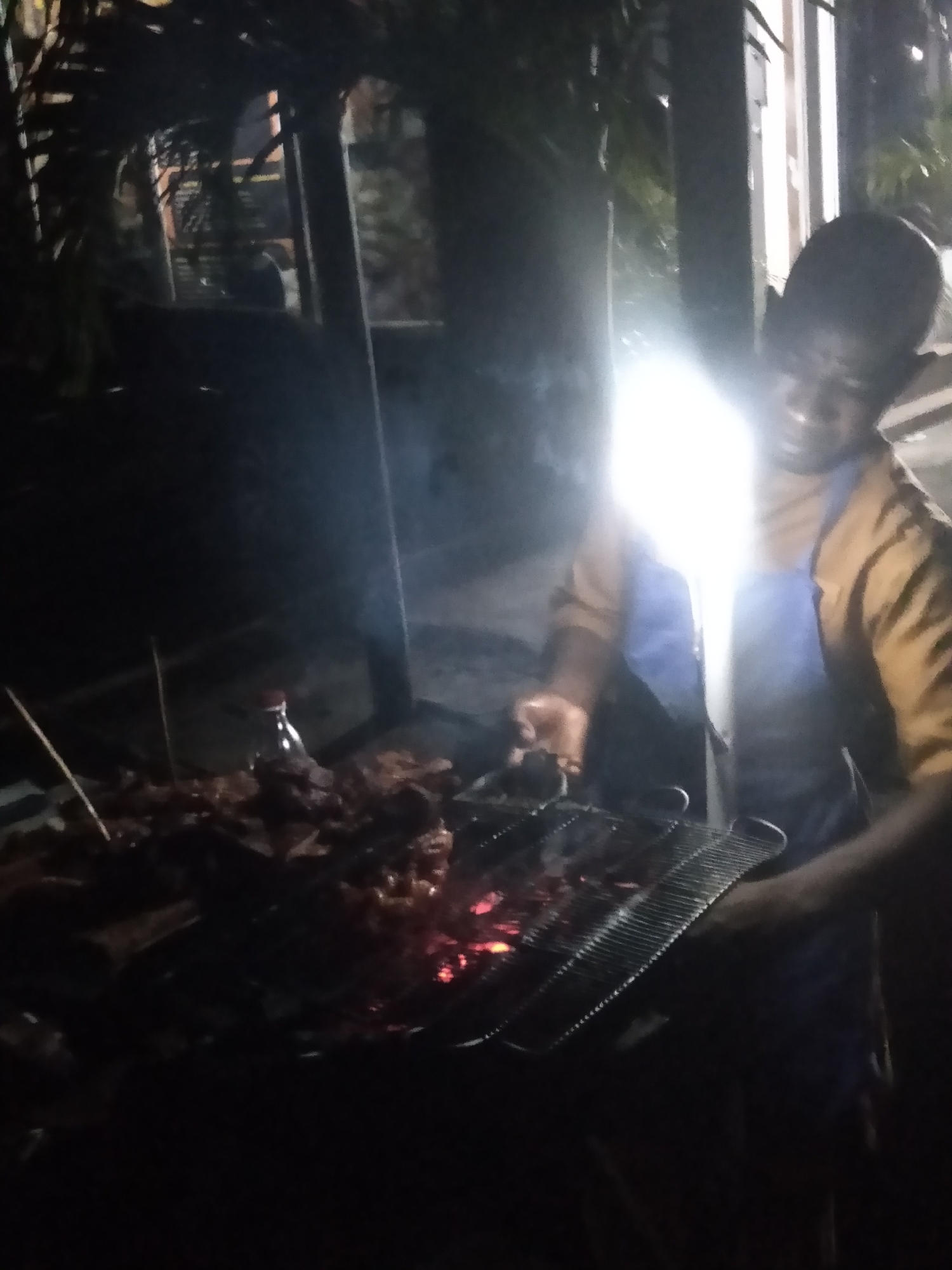 The image captures a nighttime outdoor scene in Shangisha, Nigeria, centered around a person operating a barbecue grill. The primary subject is a person, likely male, visible on the right side of the frame. They are wearing a mustard-colored long-sleeved shirt and a blue apron. Their face is partially obscured by a bright, direct light source, possibly a camera flash or a strong lamp, which creates significant glare across the upper right portion of the image.
The person is actively engaged in barbecuing, holding a utensil in their right hand, seemingly tending to the meat on the grill. The grill is visible in the lower center, with glowing red embers providing heat. Various pieces of meat, some appearing to be on skewers, are placed on the grill and piled to its left. A small, clear bottle with a red cap is also visible near the piled meat.
The setting is dark, suggesting it is late evening or night. The background is mostly obscured by shadow and the strong artificial light, but faint outlines of what appear to be trees, foliage, and possibly structural elements can be discerned, indicating an outdoor location. Smoke can be faintly seen rising from the grill. No discernible text is visible anywhere in the image. The overall impression is one of a focused individual preparing food, likely for an informal gathering, under challenging lighting conditions.