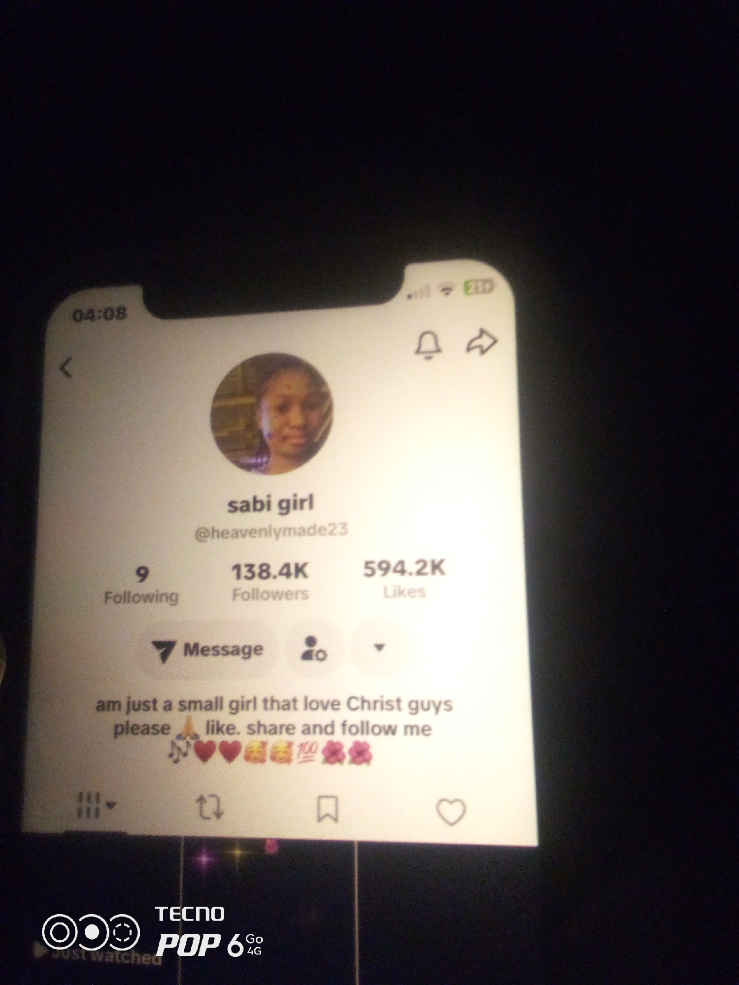 This image displays a screenshot of a social media profile, likely TikTok given the interface elements and content. The profile belongs to a user named "sabi girl" with the handle "@heavenlymade23." The profile shows statistics: 9 Following, 138.4K Followers, and 594.2K Likes. Below these statistics, there is a "Message" button and an icon representing followers. The profile's bio reads: "am just a small girl that love Christ guys please 🙏 like. share and follow me." The bio is accompanied by various emojis, including musical notes, hearts, praying hands, a fire emoji, a hundred emoji, and flower emojis. At the top of the screenshot, the time "04:08" is visible, along with cellular signal and battery indicators. The bottom of the image shows a watermark for "TECNO POP 6 Go 4G." The image itself is a photograph taken of a screen, with a dark, out-of-focus background. There is no discernible location within the city of Howayi, Nigeria, as this is a digital screenshot.