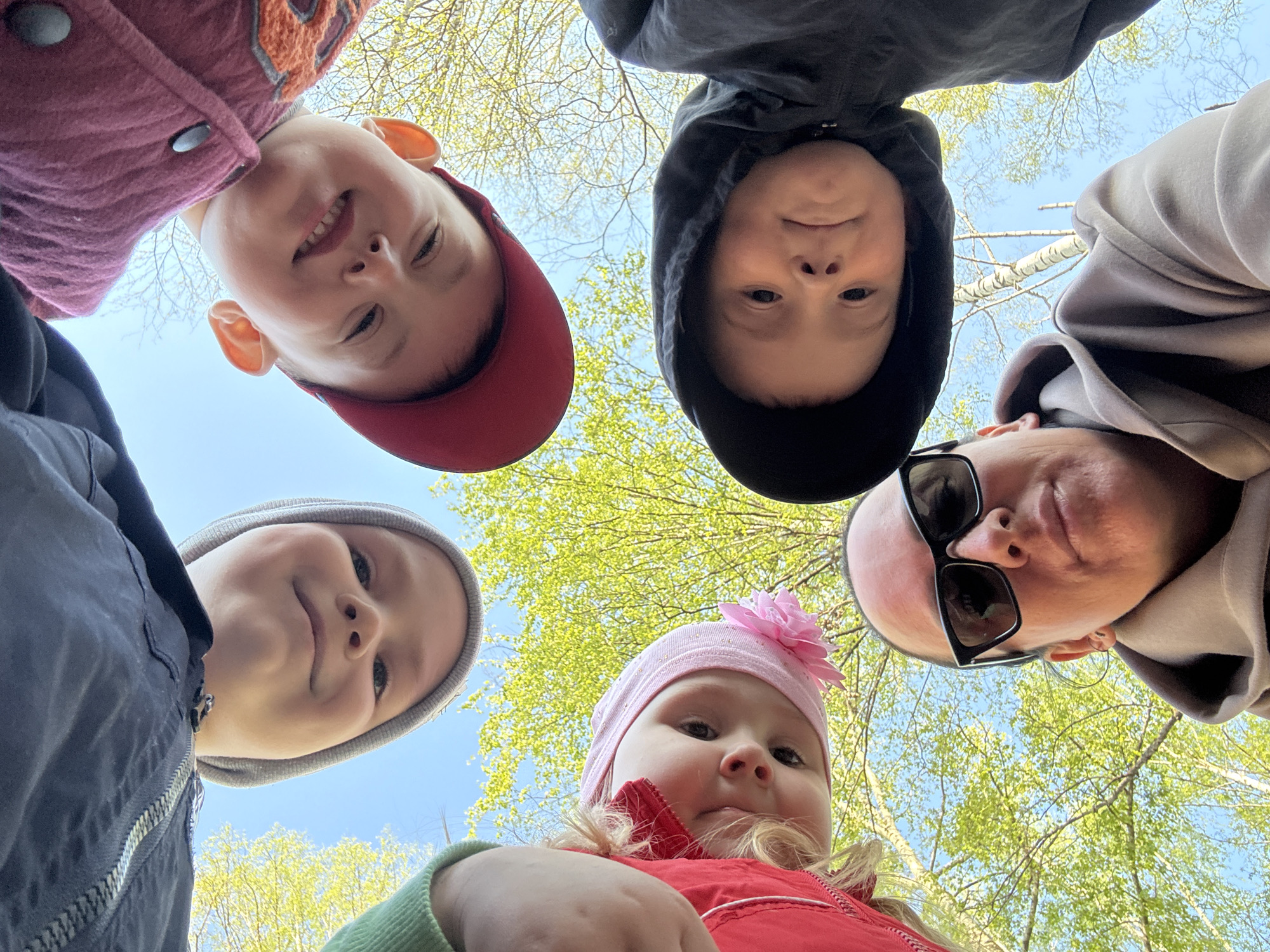 Five individuals are closely gathered, looking down at the camera. A child wearing a red cap and maroon jacket is positioned at the top-left, smiling. Next to them, a child in a black hoodie faces the camera. To the right is an adult wearing sunglasses and a light-colored hoodie. At the bottom-left, there's a child in a grey hat and blue jacket. In the bottom center, a baby wearing a pink hat with a flower and a red jacket looks up. The background shows green trees and a blue sky. The location is likely Chaikovsky, Russia, based on prior knowledge. The scene depicts a group of people, likely family, taking a photo outdoors.