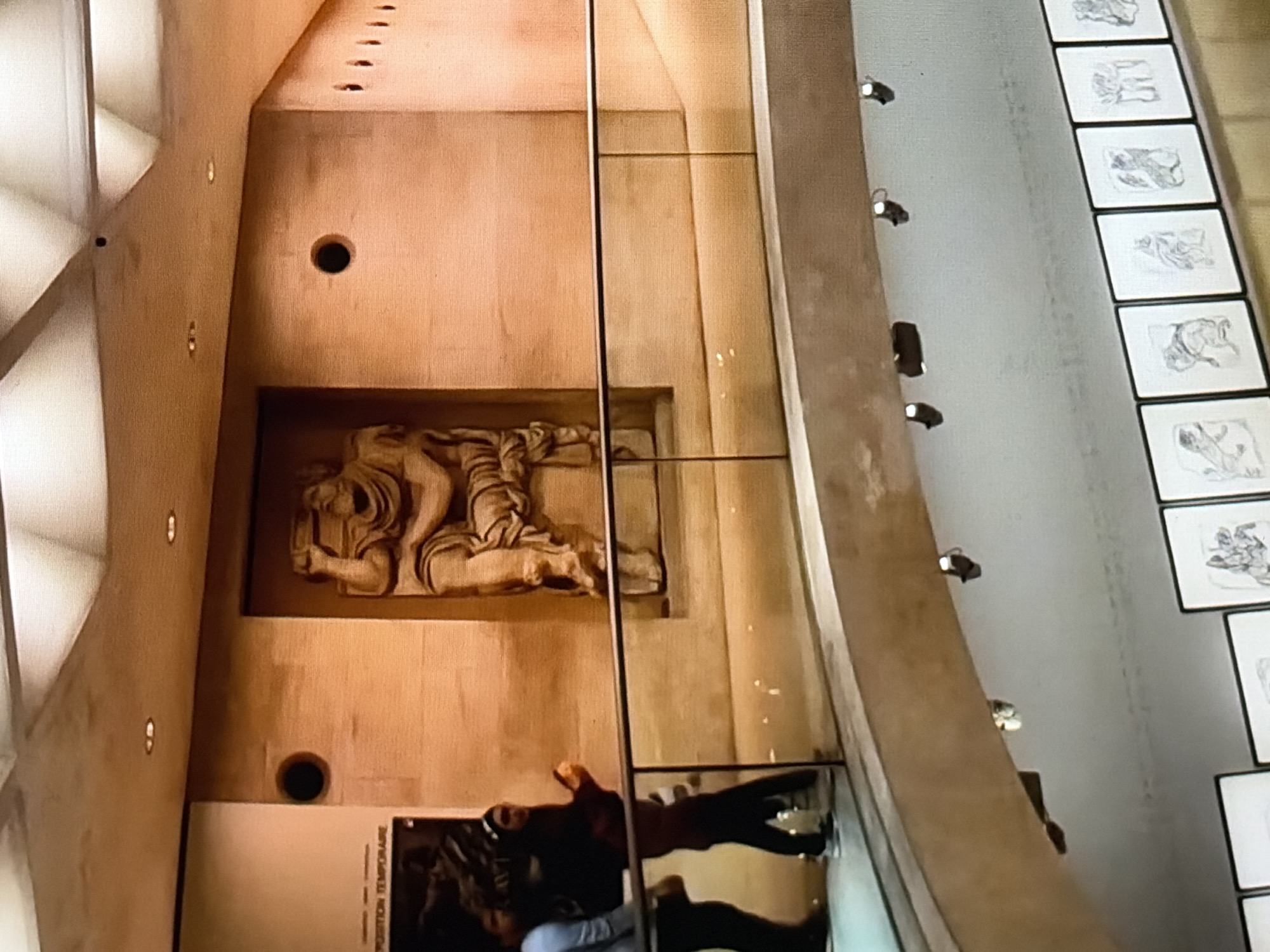 An indoor museum setting features a carved stone figure seated on a chair, holding a large object above its head, against a textured wooden wall. A transparent railing with a black top edge separates this display from a lower level. Two individuals, one in a blue shirt and the other in a dark top and pants, stand near the railing. A poster is visible on the left wall, displaying the text "EXPOSITION TEMPORAIRE." The ceiling is a series of illuminated geometric panels. Below the railing, a row of framed artworks, likely drawings, is displayed along a grey wall. The overall lighting is warm and diffused.