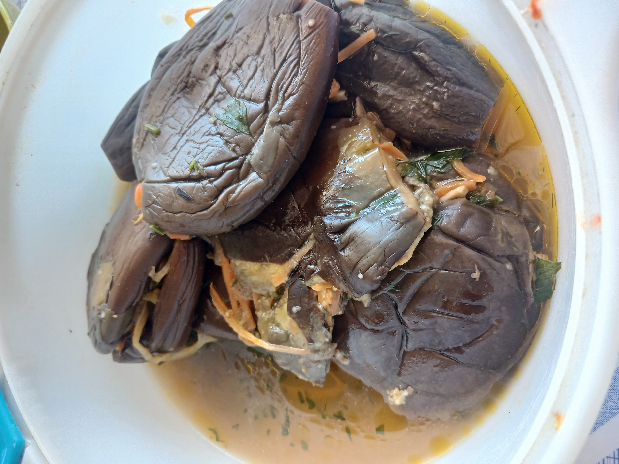 A close-up, overhead shot shows several cooked eggplants submerged in a light-colored broth within a white bowl. The eggplants are dark purple and appear to be stuffed with a filling that includes shredded orange vegetables and green herbs. The broth is slightly oily and has visible specks of herbs throughout. The bowl is white and rests on a surface that is partially visible, appearing to be a white and blue checkered tablecloth. The lighting suggests an outdoor setting during the daytime. The scene is devoid of any people or specific landmarks. The location of Hojamiresen, Turkmenistan is noted in the prompt but there are no visual cues in the image to confirm this location.