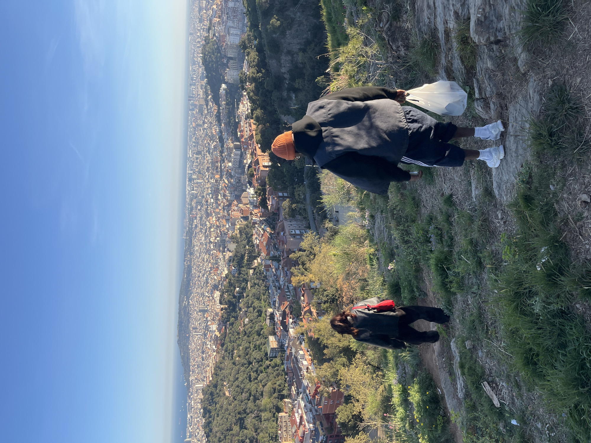 The image presents a view of Barcelona, Spain, from an elevated position. A person wearing a black jacket, orange beanie, black shorts with white stripes, and white shoes is standing on the edge of the incline, holding a white plastic bag. Another person is walking on the path to the side wearing a black jacket and red backpack. The city sprawls out below with a mix of residential and commercial buildings, interspersed with green spaces. The sky is a clear blue, transitioning to a lighter haze near the horizon where it meets the Mediterranean Sea. Vegetation, including grasses and shrubs, is visible on the incline.