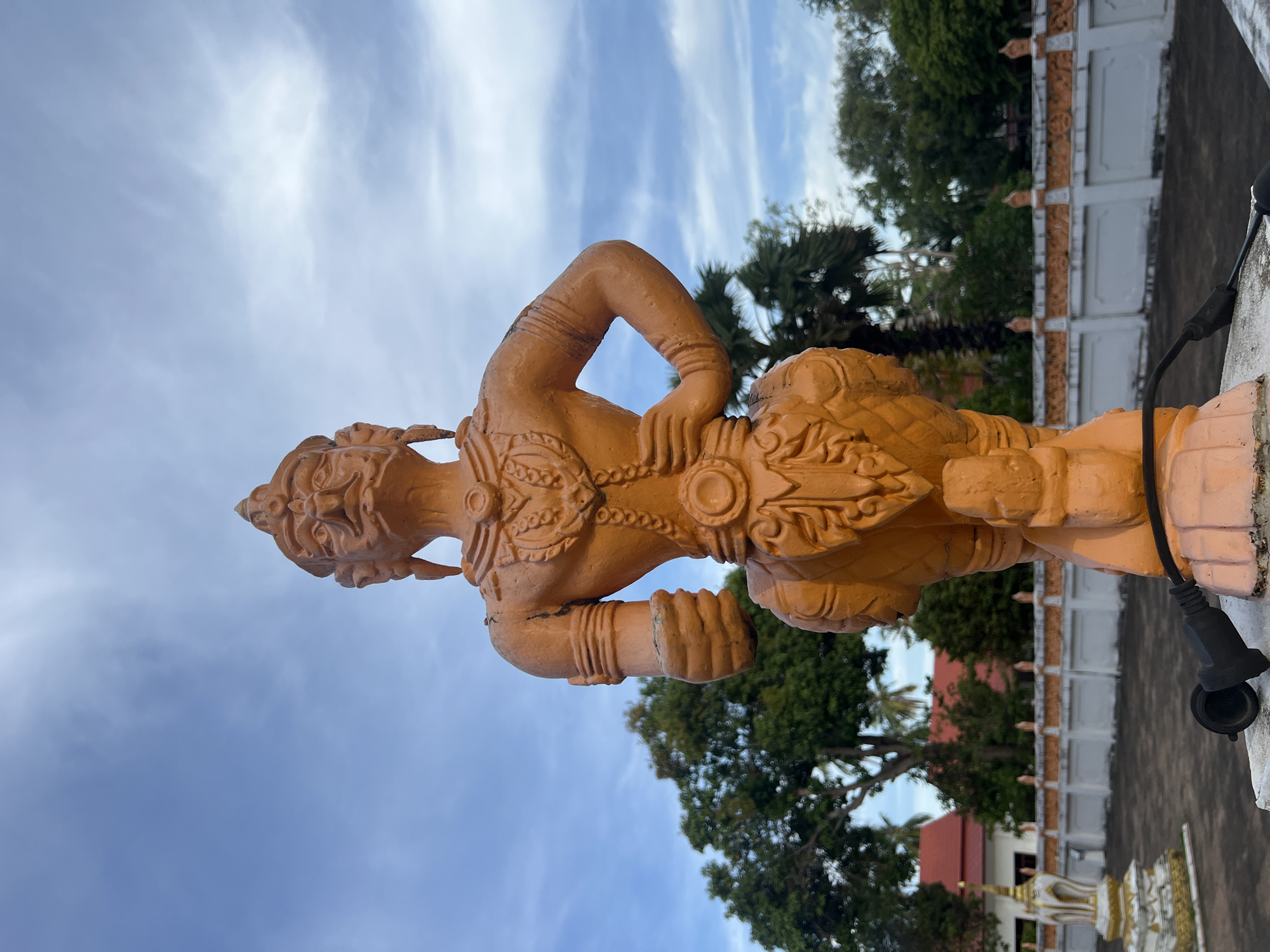 An orange-toned statue of a multi-faced mythical being stands prominently against a bright blue sky with scattered white clouds. The statue, which appears to be made of concrete or plaster, has intricate details in its headdress, chest adornments, and lower body. Its arms are positioned with one hand on its hip and the other bent at the elbow. In the background, lush green trees and palm trees are visible, along with white and orange decorative walls and buildings with red roofs, suggesting a temple or a traditional Thai architectural setting. A black electrical cord and plug are visible in the foreground, resting on the base of the statue. The overall scene appears to be outdoors in a tropical environment.