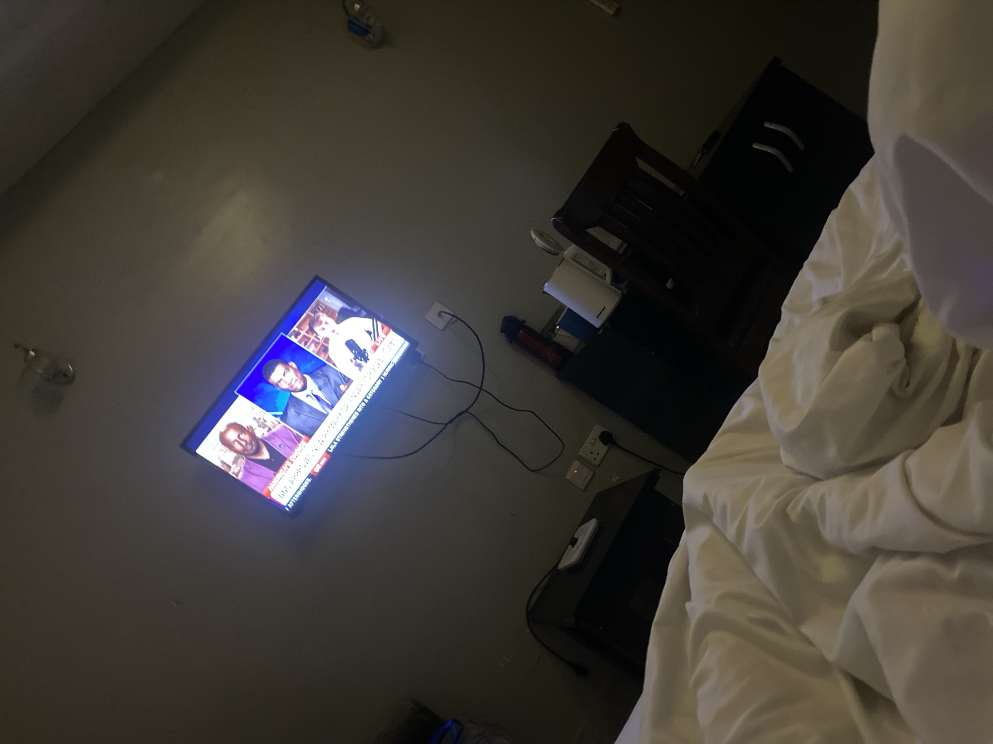 The image depicts a dimly lit bedroom scene, likely in a hotel room in Barikin Jirga Summa, Nigeria. A television screen mounted on the wall displays a news broadcast featuring three men. The room is furnished with a dark wooden chair and a small cabinet. On the cabinet, there are several items, including a white electric kettle, a red water bottle, and an electrical outlet with switches. In the foreground, a rumpled white duvet covers a bed, suggesting the viewer is lying in bed. The overall ambiance is that of a quiet, private space in the evening or night.