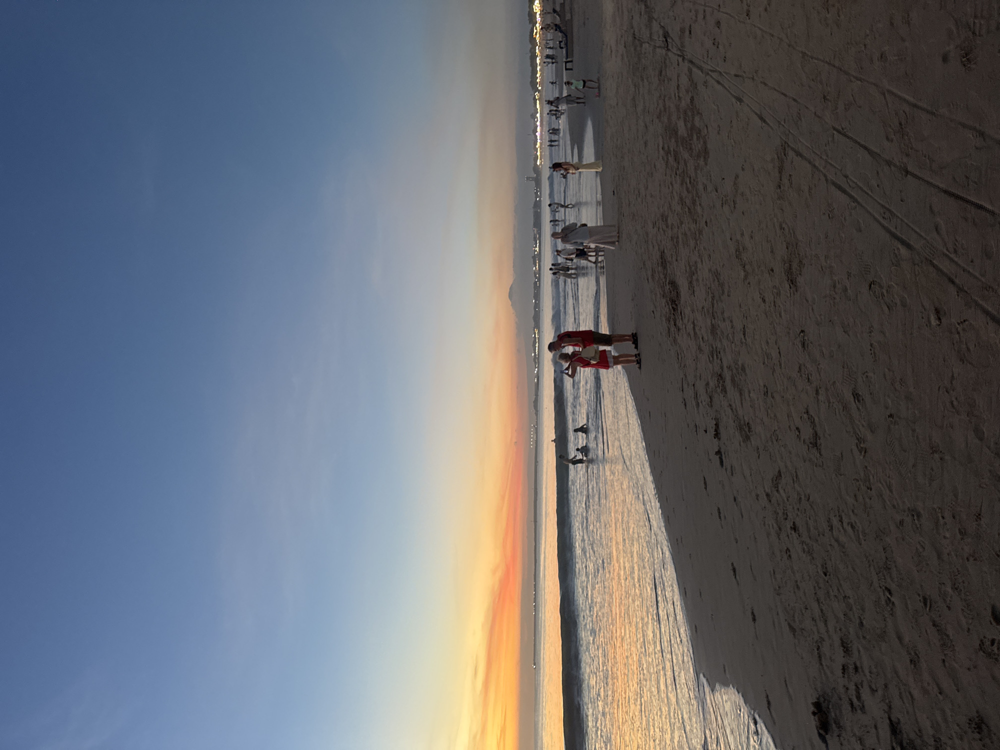 The image depicts a beach at sunset, likely in Kuta Selatan, Indonesia. The sky transitions from blue to orange near the horizon, where the sun is setting. The beach is sandy and slopes down to the water's edge. Several people are present, some standing near the water and others further up the beach. There are visible footprints and tire tracks in the sand. The water reflects the colors of the sunset. In the distance, there are city lights.