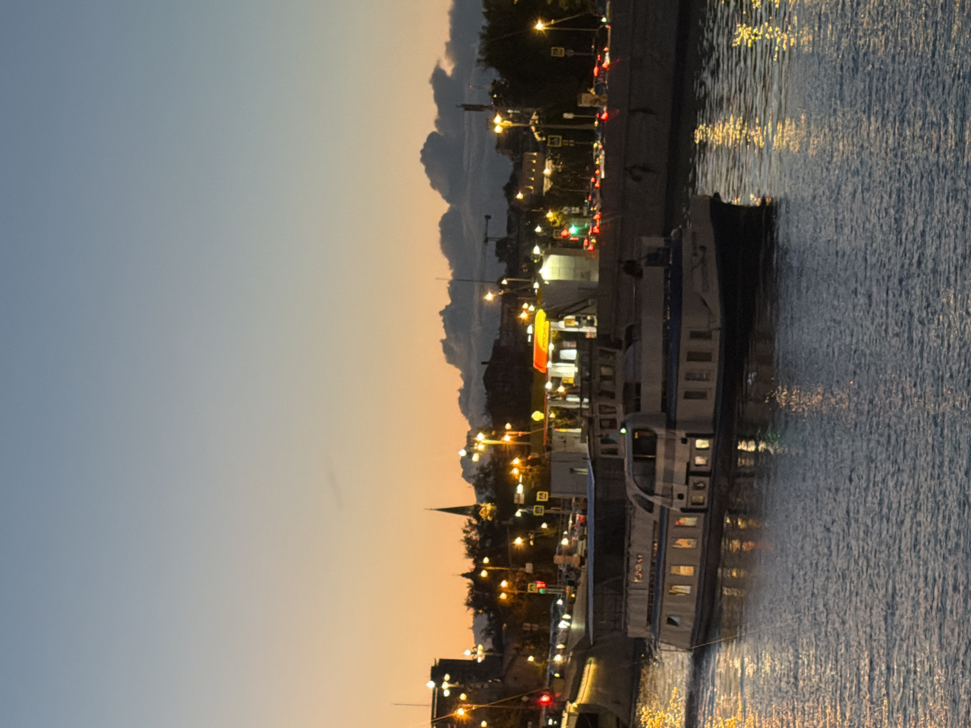 The image captures a twilight scene over a body of water, likely a river or canal, in Moscow, Russia. A white and blue passenger boat is the main subject, cruising from left to right across the frame. The boat has multiple windows illuminated from within, and its reflection shimmers on the water's surface, along with the reflections of city lights.
In the background, the Moscow skyline is visible. The lights of buildings, street lamps, and traffic can be seen. The sky transitions from a warm orange hue near the horizon to a cooler blue overhead, indicative of sunset or twilight. A silhouette of a church steeple is prominent against the sky. The water appears choppy, with ripples and reflections distorting the light sources. The overall impression is one of a bustling city at dusk, with urban life continuing on the water and along the embankment.