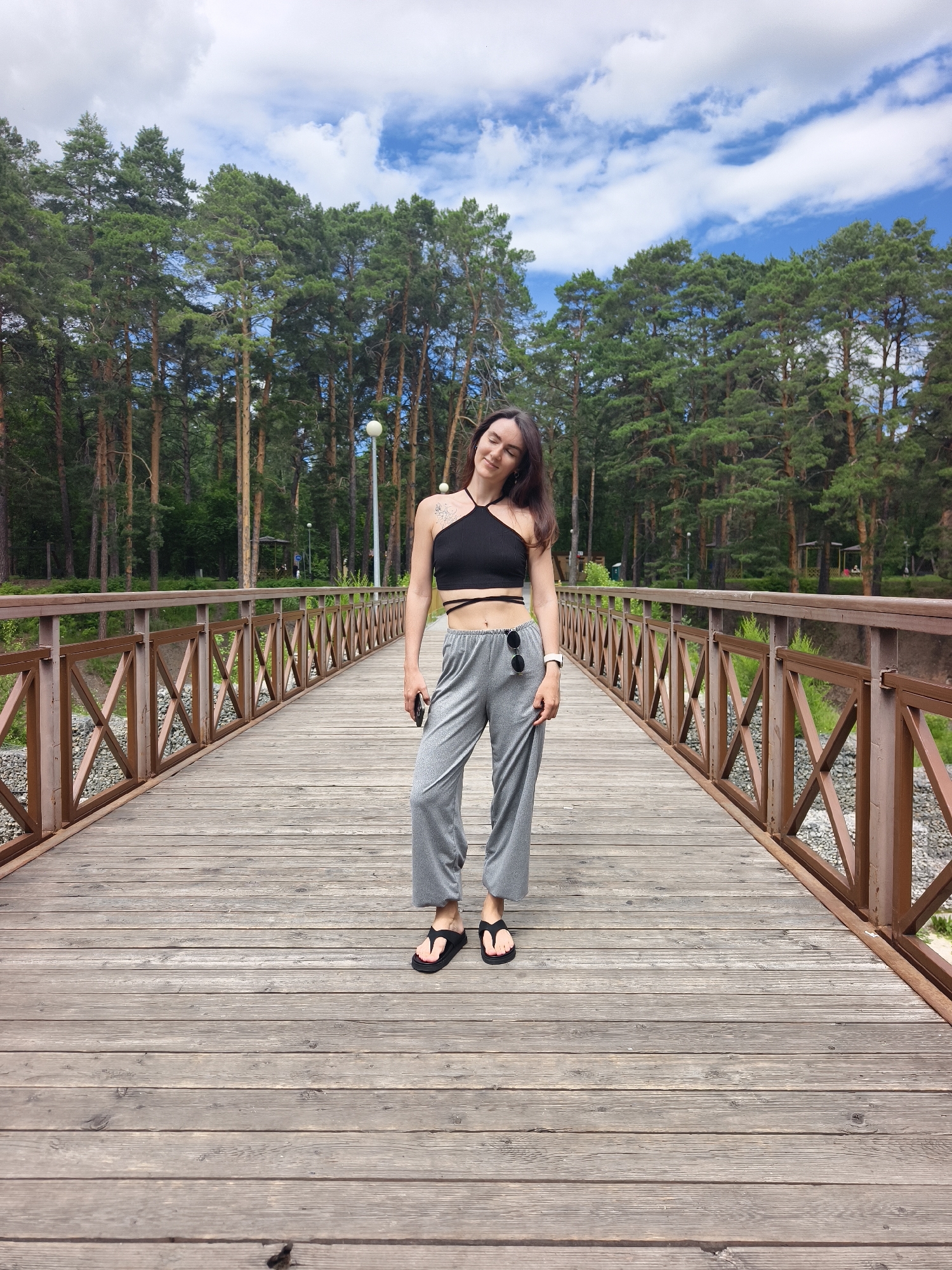 A young woman stands on a wooden bridge in a park. She is wearing a black crop top, gray wide-leg pants, and black flip-flops. She has dark hair and is looking down with her eyes closed. Sunglasses are tucked into her pants pocket, and a smartwatch is on her left wrist. The bridge has a brown wooden railing with a crisscross pattern. Behind her, tall pine trees line the edge of a body of water. The sky is blue with white clouds. The location is the Arboretum in Tyumen, Russia.