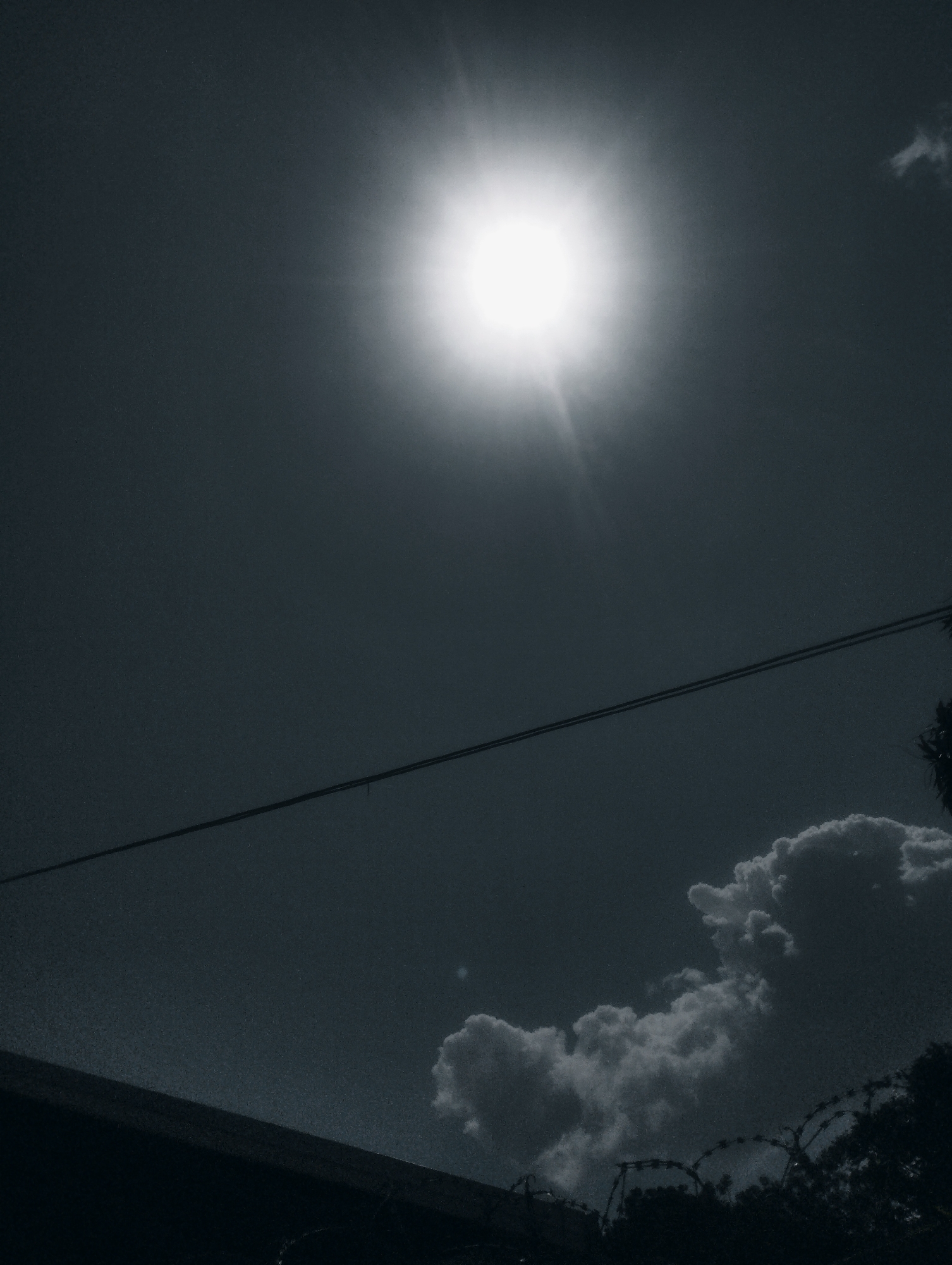 The image is a high-angle, monochromatic shot taken outdoors, likely during the daytime in Jos, Nigeria, as suggested by the presence of the sun and a dark, silhouetted foreground that could be a building or structure. The sun is the dominant element in the upper portion of the frame, appearing as a bright, blown-out circle with visible rays, indicating it is high in the sky. Two parallel power lines stretch horizontally across the mid-section of the image. Below these lines, in the lower right quadrant, are several fluffy, white clouds against a dark sky. The bottom left of the image features a dark, angled shape, possibly a rooftop or wall, and at the very bottom, barbed wire is visible on top of some foliage. The overall mood is stark and high-contrast due to the monochromatic tone and the bright sun. There are no people visible in the frame. The environment suggests an urban or semi-urban setting with infrastructure like power lines and fencing, combined with natural elements like the sky and clouds. The image has a grainy texture.