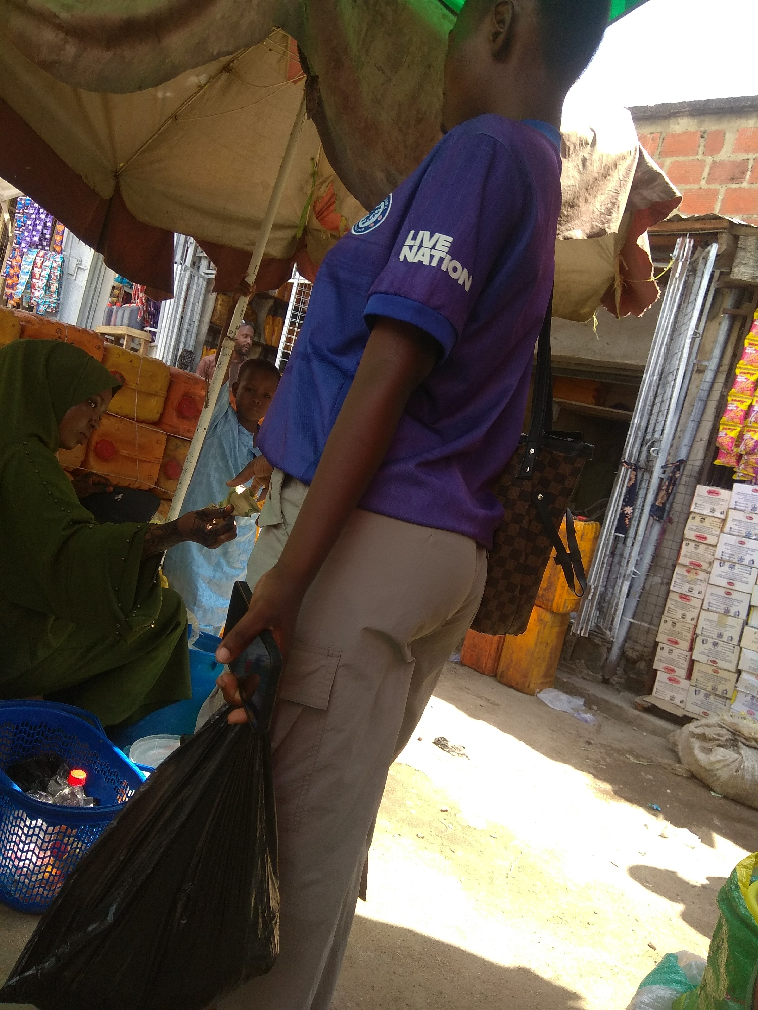 In a bustling outdoor market in Jalingo, Nigeria, a person wearing a purple t-shirt with "LIVE NATION" printed on it is seen from the side, holding a black plastic bag. The person is also carrying a brown checkered shoulder bag. In the foreground, a blue plastic basket contains items including a water bottle with a red cap. To the left, a woman wearing a green hijab is handing a banknote to a child. The background shows stacks of yellow and orange containers, shelves lined with packaged goods, and a brick building. The scene is illuminated by sunlight casting shadows on the dusty ground.