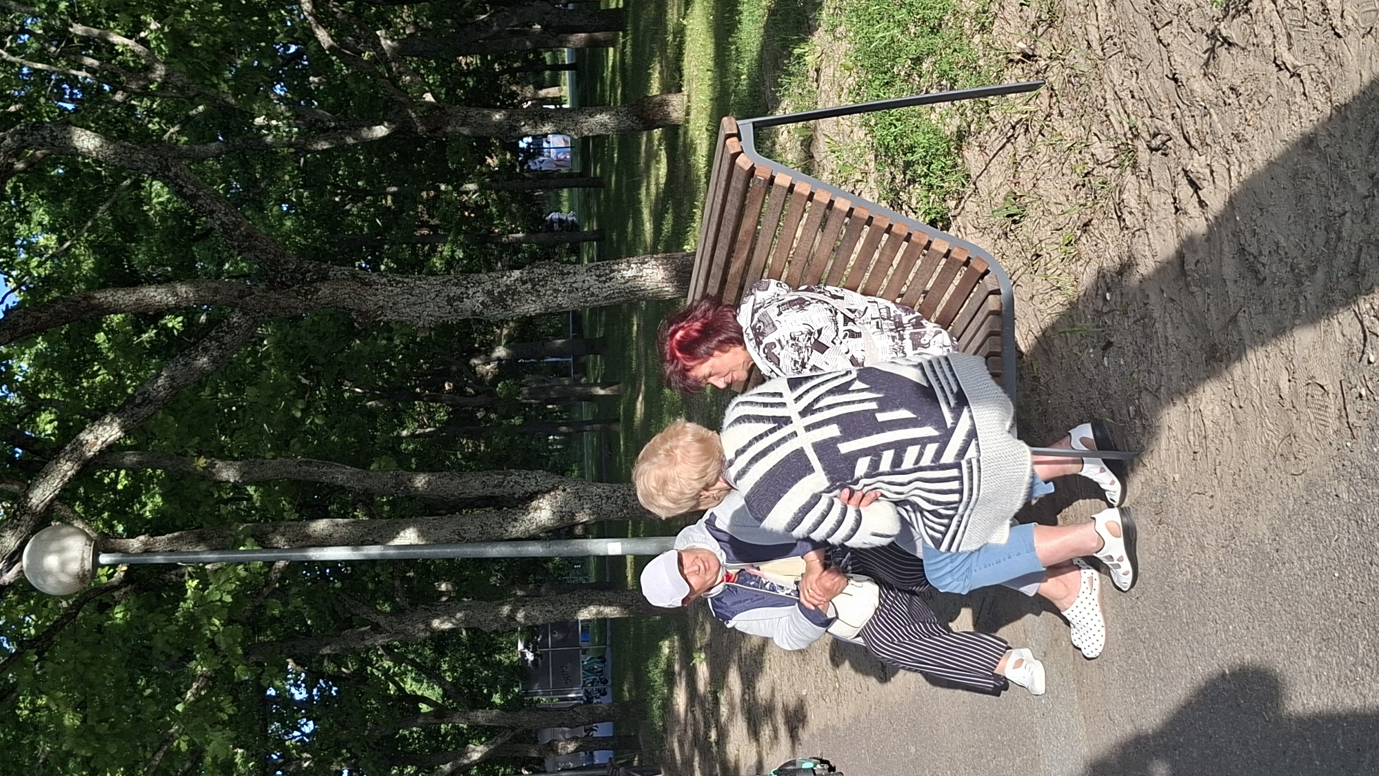 In the city of Tallinn, Estonia, three women are seated on a park bench under a canopy of green trees. The woman on the left, wearing a white cap, blue and white jacket, and striped pants, is looking at the woman in the middle. The woman in the middle, with blonde hair, is wearing a black and white patterned sweater and is facing away from the camera, towards the woman on the right. The woman on the right, with red-tinted hair, is wearing a black and white patterned jacket and is looking at the woman in the middle. A lamppost stands behind them, and the ground is a mixture of gravel and dirt, with scattered patches of grass. The overall scene depicts a casual conversation among friends in a park setting.