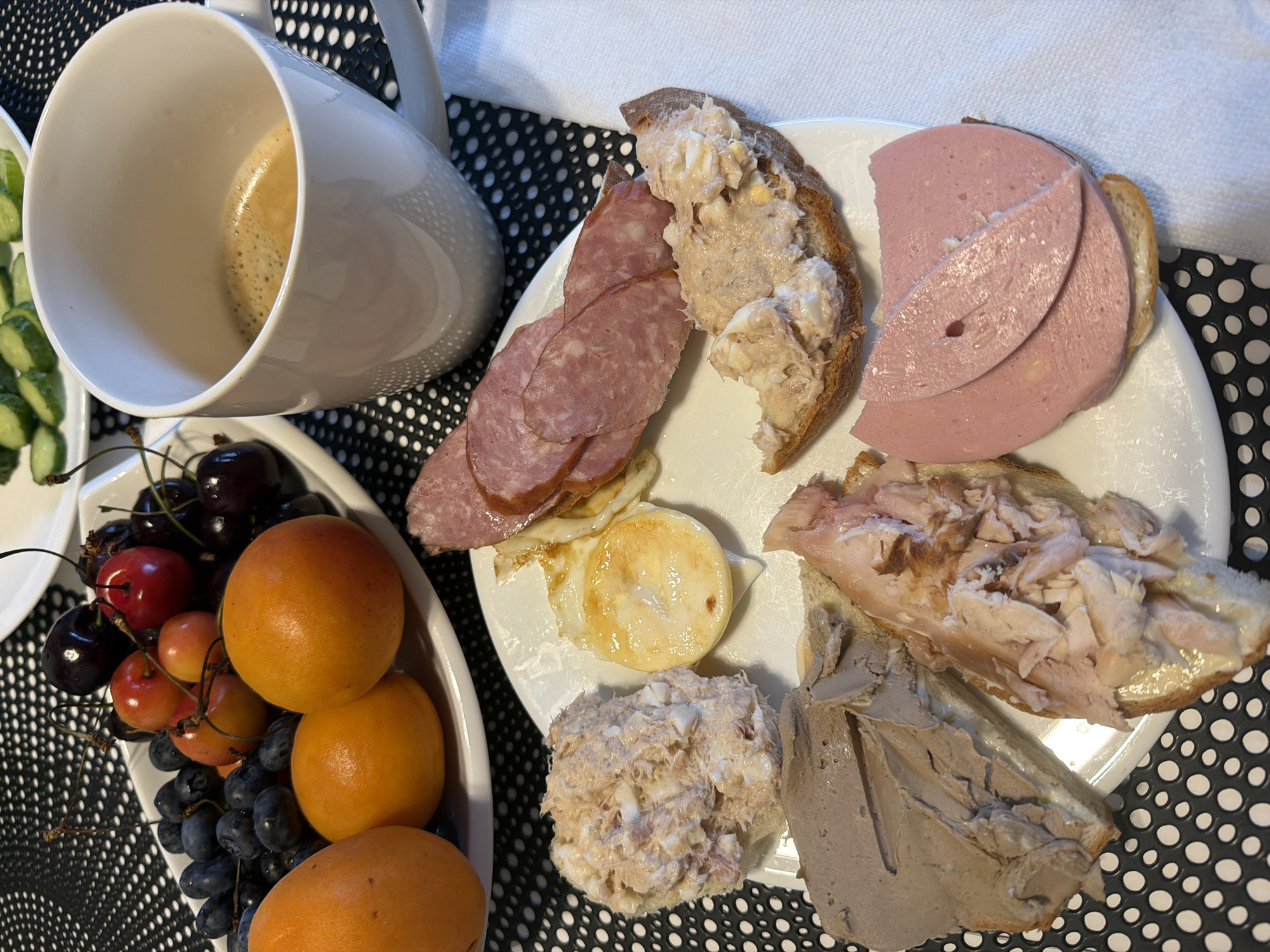 The image displays a breakfast spread laid out on a black and white speckled tablecloth, likely in a home environment in Baloži, Latvia. The breakfast includes a white plate filled with various food items. On the plate, there are open-faced sandwiches topped with different spreads and meats. These include a sandwich with what appears to be pate, a sandwich with shredded chicken, a sandwich with a creamy mixture possibly containing tuna and egg, and a sandwich topped with sliced bologna. Adjacent to these sandwiches is a fried egg on top of another slice of bread. To the left of the plate of sandwiches is a white oval dish filled with fruit, featuring dark grapes, red cherries, and orange apricots. A white mug filled with coffee sits to the right of the plate. In the background, a plate of sliced cucumbers is partially visible. The overall scene suggests a casual breakfast setting.