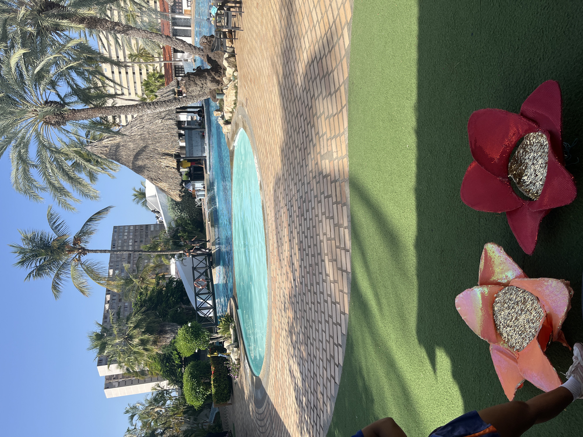 The image shows a resort swimming pool area. A large, light-colored building dominates the background, surrounded by palm trees and other tropical vegetation. A thatched-roof bar area is situated next to the pool. The pool itself is a bright blue, with a small bridge arching over one end. The pool deck is paved with light-colored bricks arranged in a circular pattern. In the foreground, two artificial flower decorations, one pink and one red, are placed on a patch of artificial grass. Both flowers are filled with birdseed. Several people are visible in and around the pool area, including some standing on the bridge and others near the bar. The sky is clear and blue, indicating a sunny day.