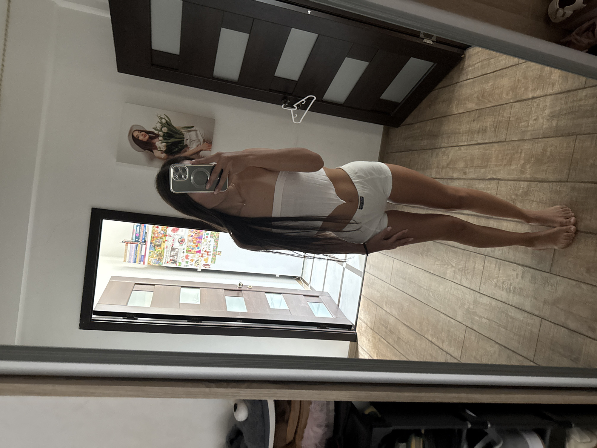 A young woman with long dark hair stands in a room, taking a selfie in a mirror. She is wearing a white bandeau top and white shorts. Her reflection shows a picture on the wall behind her of a woman holding a bouquet of tulips. The room has wooden floors and two dark wood doors, one of which is open. A shelf unit with shoes and other items is visible to the left. This image was taken in a residential interior, likely a bedroom, in Chișinău, Moldova.
