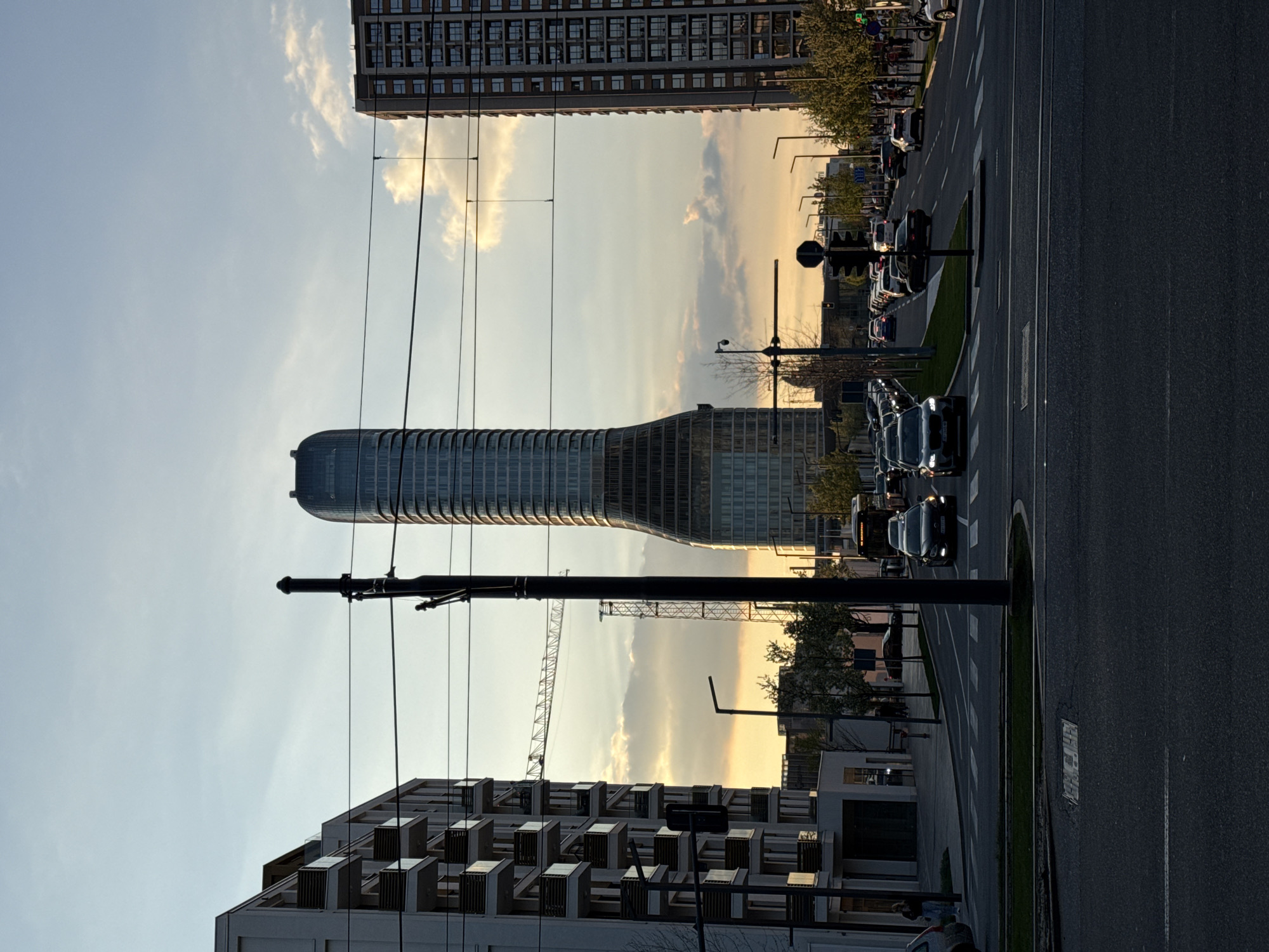 The image shows a street scene in Belgrade, Serbia, at sunset. Prominent architectural features include the distinctive silhouette of the West 65 Tower, appearing squeezed in the middle, alongside other high-rise residential and commercial buildings. A construction crane is visible in the background, suggesting ongoing urban development. The street is lined with cars, indicating moderate traffic. Utility poles and power lines cross the frame. The sky is filled with soft hues of orange and yellow, indicative of the setting sun. A sidewalk is visible, with little to no pedestrian traffic in sight. The image is oriented vertically, with buildings running along the top and bottom and the street running along the right edge.