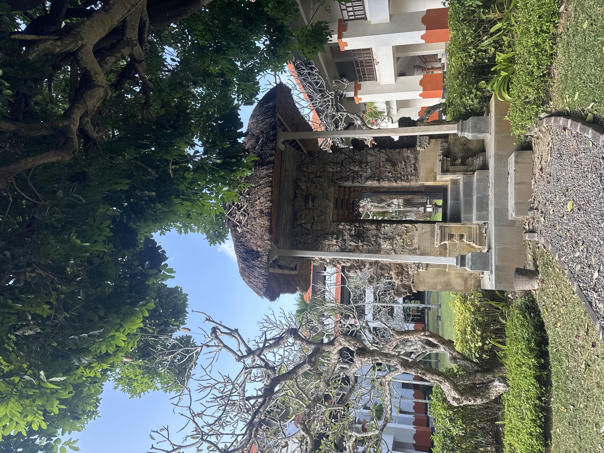 A sunlit, outdoor scene captures a small, ornate stone structure in Kuta Selatan, Indonesia. The structure, likely an altar or temple entrance, features intricate carvings and a thatched roof, supported by thick pillars. It is approached by a set of stone steps. To the left, a bare, twisted tree with delicate branches stands in front of a low-rise hotel building. The hotel features white walls with orange accents around balconies and windows, and a red tiled roof. Lush green bushes and manicured grass surround the stone structure and the hotel, suggesting a well-maintained resort or garden setting. The sky above is clear and blue.
