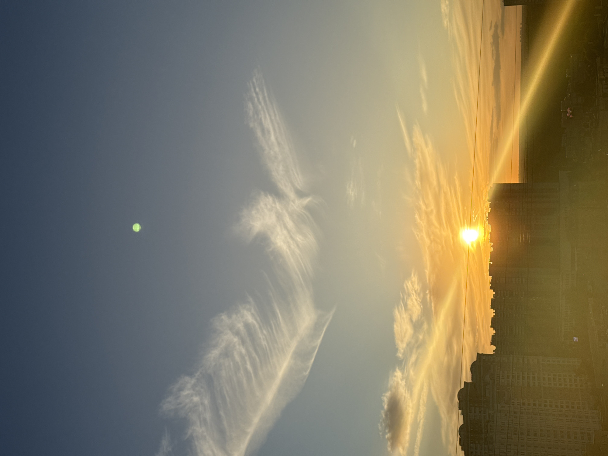 The image depicts a sunset over the city of Khimki, Russia. The sky is a gradient of deep blue at the top, transitioning to lighter blues and then to a warm orange and yellow glow near the horizon. Wispy white clouds are scattered across the sky, with some appearing more dense and others thin and feathery. A distinct, bright green circular object is visible in the upper portion of the sky.
In the foreground, the silhouettes of several large apartment buildings dominate the lower part of the frame. These buildings are modern, with multiple floors and visible windows, characteristic of urban housing complexes. The setting sun is a bright, intense orb positioned near the right side of the frame, casting a strong golden light that creates a lens flare effect, with a prominent diagonal ray of light extending across the lower right quadrant of the image. The sun's rays illuminate the sky and create highlights on the clouds and the tops of the buildings.
The lower portion of the image shows the urban landscape extending into the distance, with more buildings and structures visible. The overall impression is of a serene evening scene in a densely populated urban environment, with the dramatic colors of the sunset adding a striking visual element to the cityscape. No people or specific actions are discernible.