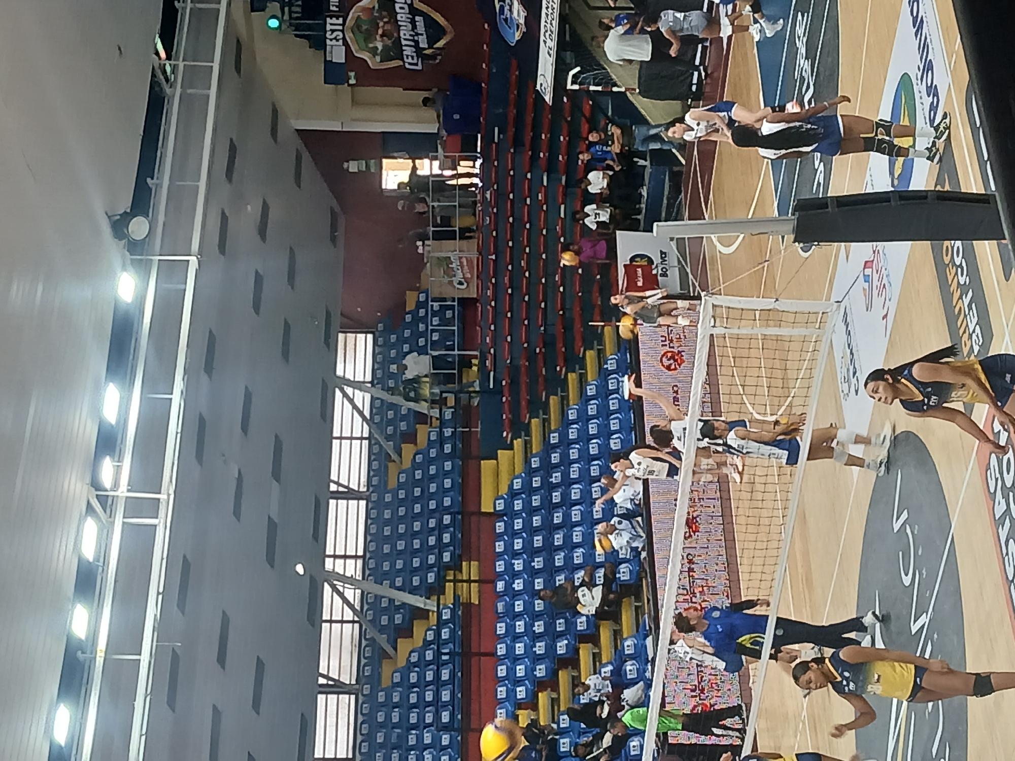 A volleyball match is in progress at an indoor stadium in Ciudad Guayana, Venezuela. Two teams, one in blue and white uniforms, and the other in yellow and dark blue, compete on a wooden court divided by a net. The stands, filled with spectators, are primarily blue and red with some yellow sections. Banners and advertisements are visible on the court and in the stands, including the word "Bolivar" and a logo for "Central de Bolívar." Bright overhead lighting illuminates the scene.