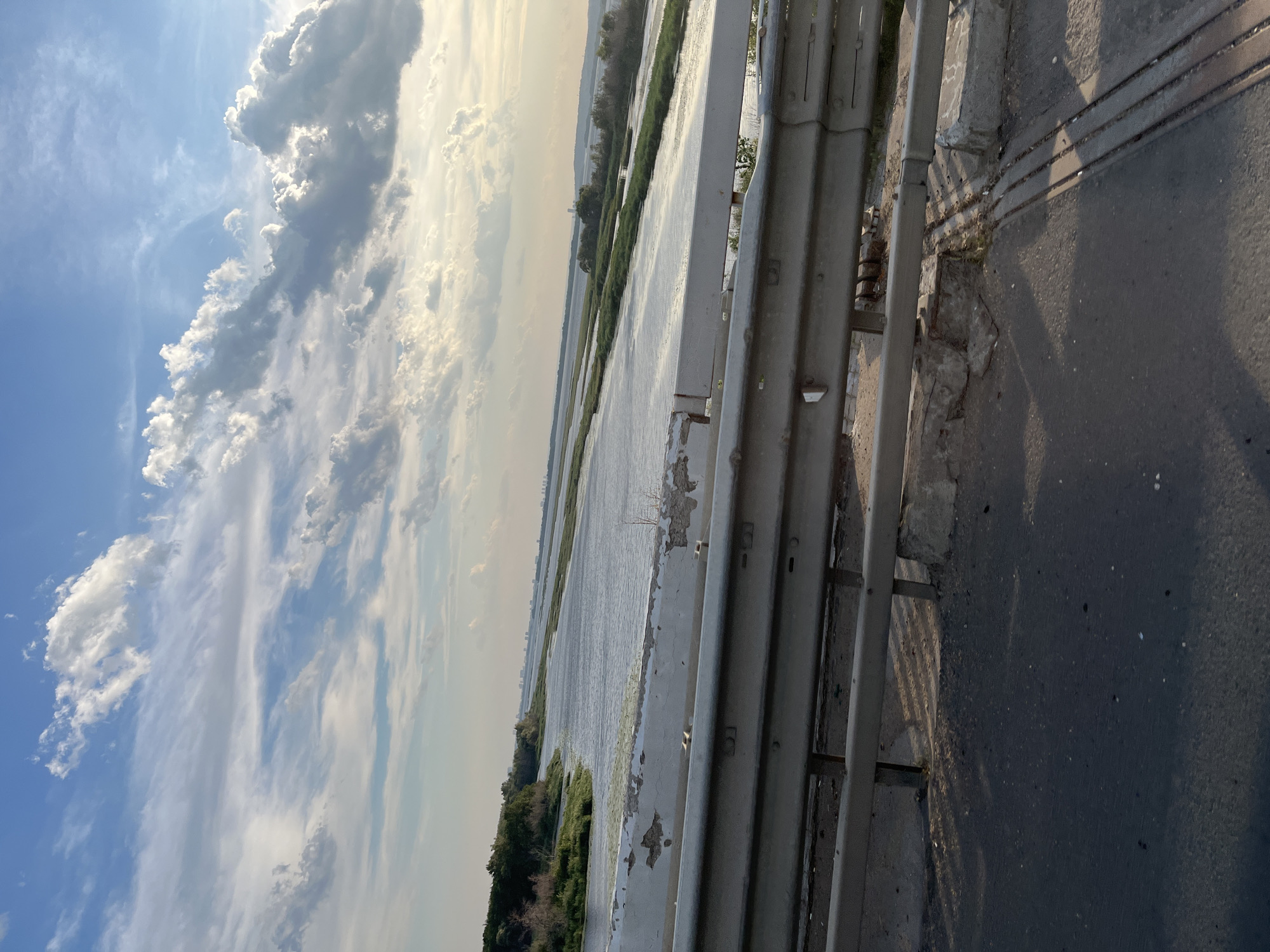 The image captures a wide river with several green islands of vegetation, stretching towards a distant city skyline under a cloudy blue sky. In the foreground, a weathered metal guardrail runs along a paved road, presumably a bridge, offering a glimpse of the road surface and concrete supports. The setting appears to be the Volga River in Shumeika, Russia, given the presence of the characteristic landscape and the distant urban silhouette. There are no people or vehicles visible in the frame.
