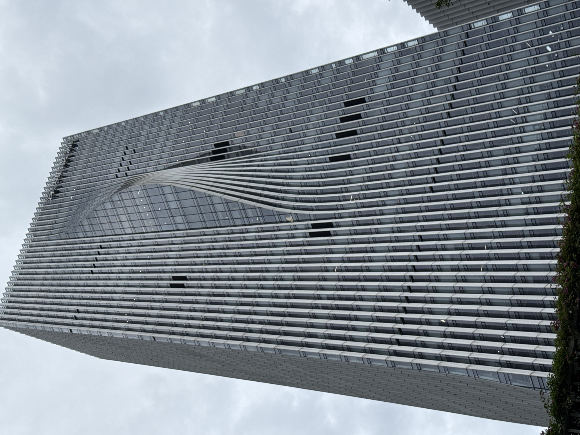 The image shows a tall, modern skyscraper with a unique twisted design in Futian District, China. The building has a glass facade with vertical silver fins that create a textured appearance. The fins are angled and appear to curve as they ascend the building, giving it a dynamic and fluid look. The sky is overcast, with grey clouds. The bottom of the image shows some greenery, suggesting a park or landscaped area at the base of the skyscraper. No people or vehicles are visible.