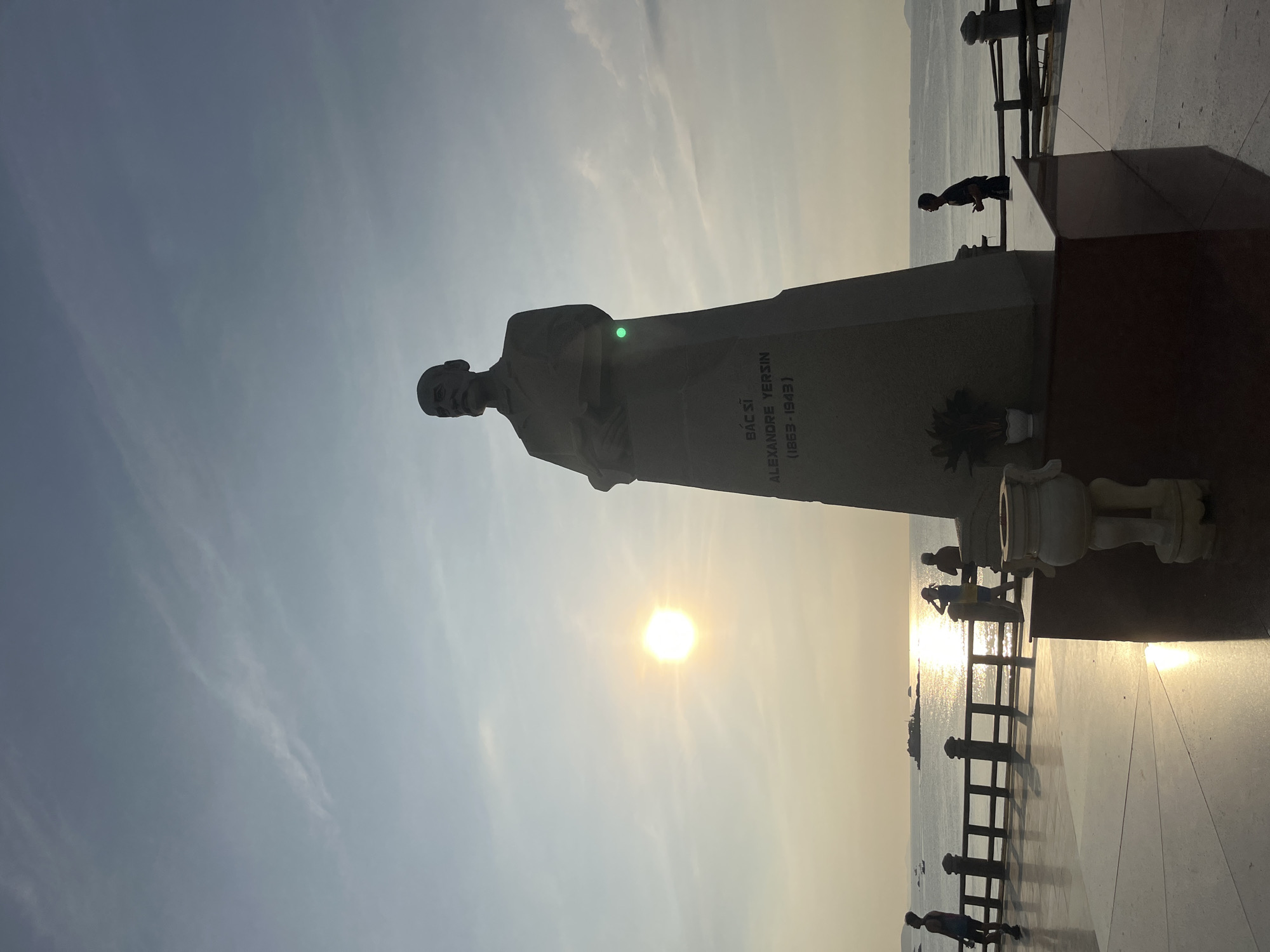 The image shows the Alexandre Yersin statue in Nha Trang, Vietnam, silhouetted against a bright sky. The sun is visible. The statue stands on a raised platform with railings along the waterfront. Several people are walking and standing near the railings.