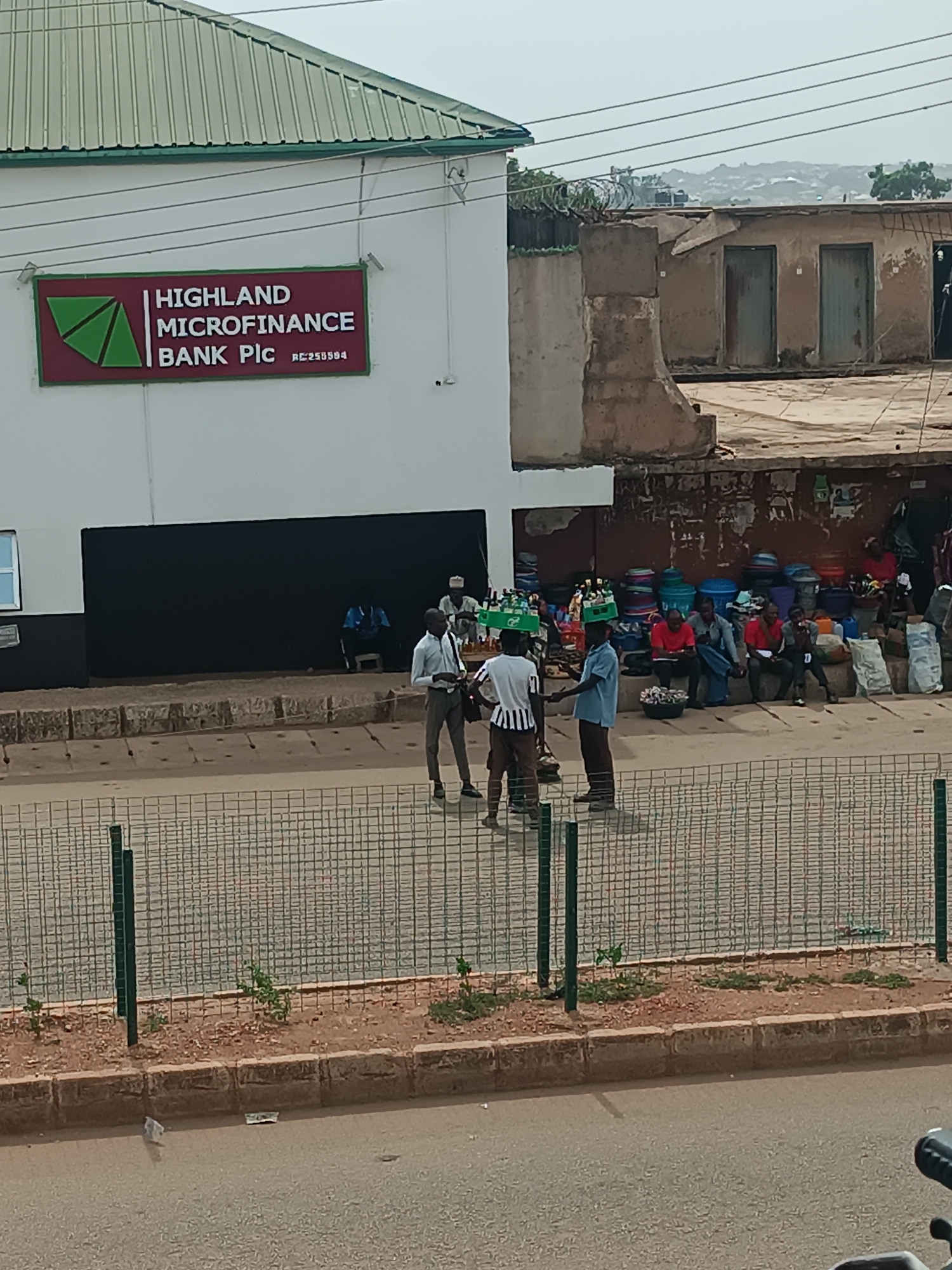 The image shows an outdoor scene in Bukuru, Nigeria, during the daytime. The primary subject is the front of a building identified as "Highland Microfinance Bank Plc" by a sign on the facade. The building has a green-trimmed roof and a large black horizontal band below the signage.
In front of the bank, there are three men engaged in a conversation. To the right, a group of people are sitting on a ledge, possibly waiting or observing the scene. Behind them, there are piles of goods, suggesting a market area.
A metal mesh fence runs along the edge of the street in the foreground. The road is visible and appears to be a light brown color.
In the background, there is another building that appears to be in a state of disrepair. Power lines stretch across the sky above the buildings.