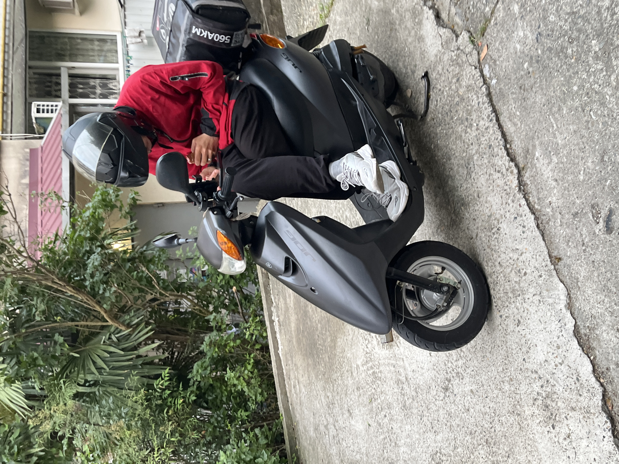 A person wearing a black helmet and a red jacket is sitting on a black scooter. The scooter has a black delivery bag on the back with the text "560AKM" printed on it. The person is holding a phone and appears to be looking at it. The scooter is parked on a concrete surface. In the background, there are lush green trees and a building with a red roof. The scene is outdoors, with soft daylight. The environment appears to be a residential area. The image does not depict the city of Sochi, Russia.
