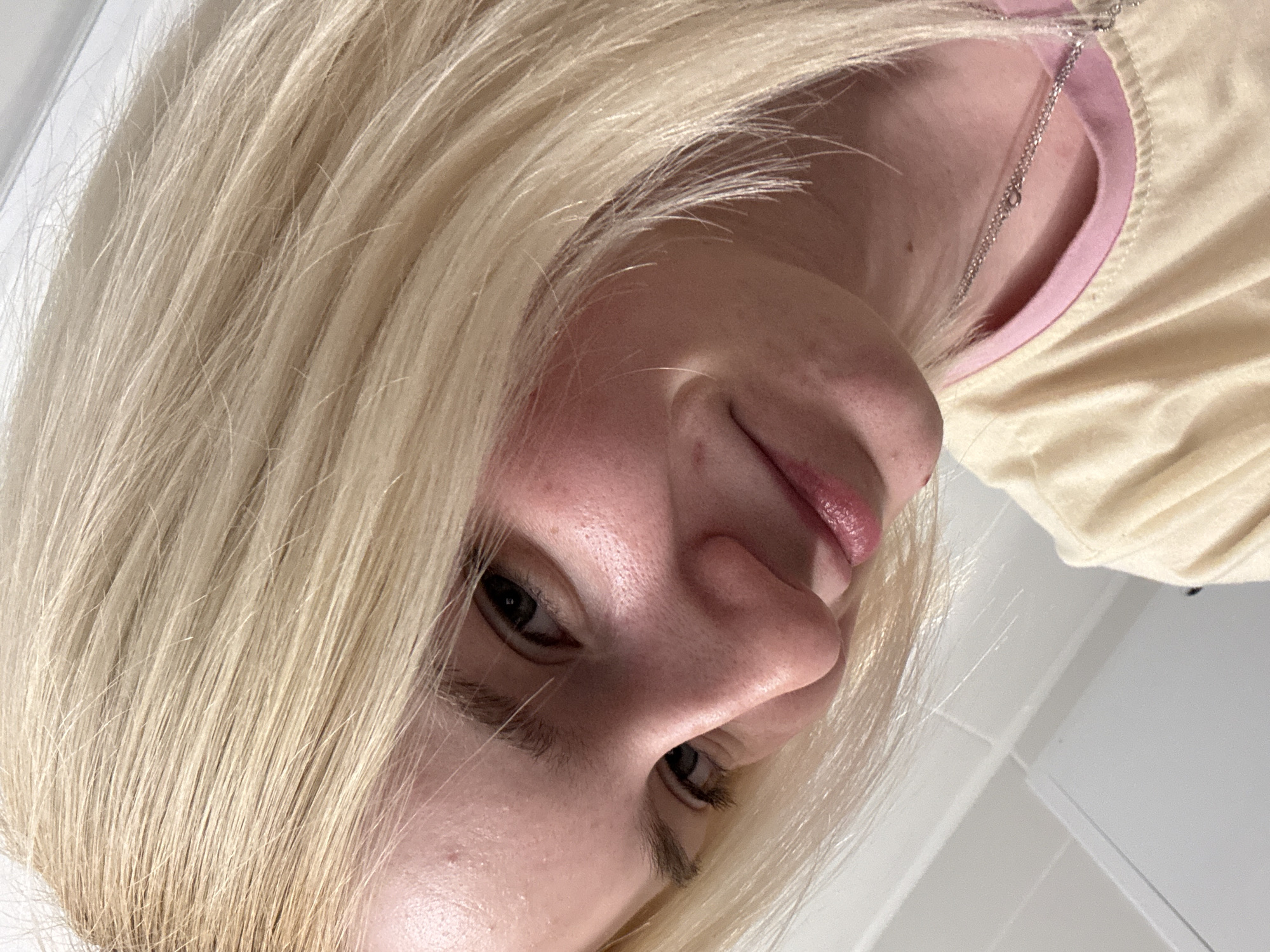 A close-up, downward-angled selfie of a light-skinned person with blonde hair is shown. Only a portion of their face is visible, including their eyes, nose, and lips. They are wearing a light yellow shirt with a pink neckline. A silver chain necklace is also visible. The background is a plain white surface, possibly a wall. There is no information available to determine the location as Vsevolozhsk, Russia.