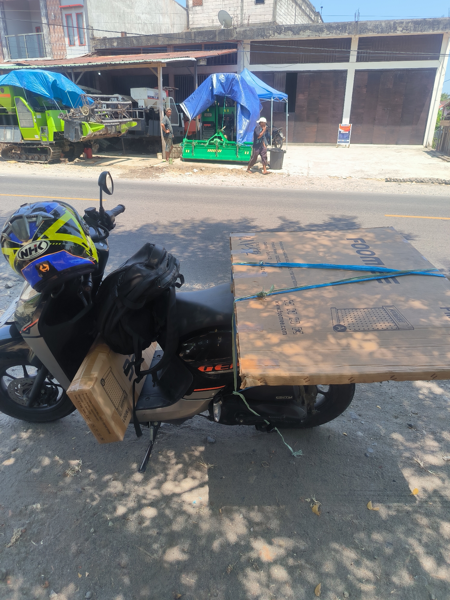 A motorcycle is parked on the side of a road in Pinrang, Indonesia, with a large, flat cardboard box strapped to its rear. The box is secured with blue and green ropes and has "FOOTME" printed on it along with a website address. A smaller cardboard box is also attached to the front of the motorcycle. On the motorcycle's seat, a black backpack and a blue and yellow NHK helmet are placed. In the background, a building with large, dark brown garage doors is visible, with a man in a white hat and dark clothing walking away from a trash can. Further back, agricultural machinery covered with blue tarpaulins is parked under a shelter. The scene is illuminated by sunlight, casting shadows on the ground.