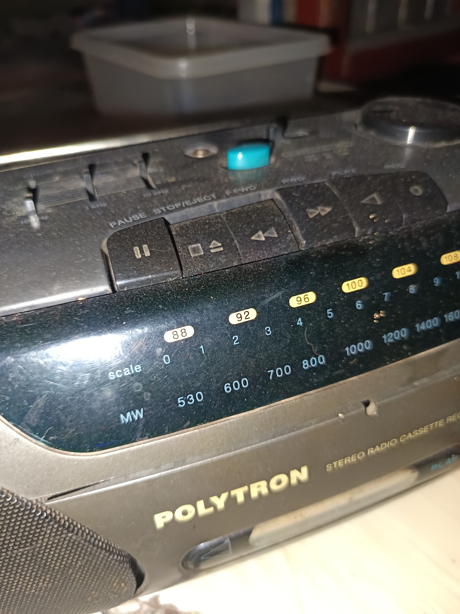 The image shows a close-up, high-angle view of a dusty, black Polytron brand stereo radio cassette recorder. The device has several buttons on its top panel, including a blue "PAUSE STOP/EJECT" button and buttons with symbols for play, rewind, and fast forward. The front panel features a tuning dial with illuminated frequency numbers such as "88", "92", "96", "100", "104", and "108" for FM, and numbers like "530", "600", "700", "800", "1000", "1200", "1400", and "1600" for MW (Medium Wave). The "MW" label is clearly visible. The overall condition of the device suggests it is an older model and has been exposed to dust and wear. The background is out of focus, showing what appears to be a light-colored plastic container and some shelves with indistinct items. The lighting is dim, creating some glare on the glossy surfaces of the radio. There are no people or specific Indonesian landmarks visible in the image, and no direct indication of the location being Sirnagalih, Indonesia, beyond what might be inferred from the brand of the device or its commonality in that region.