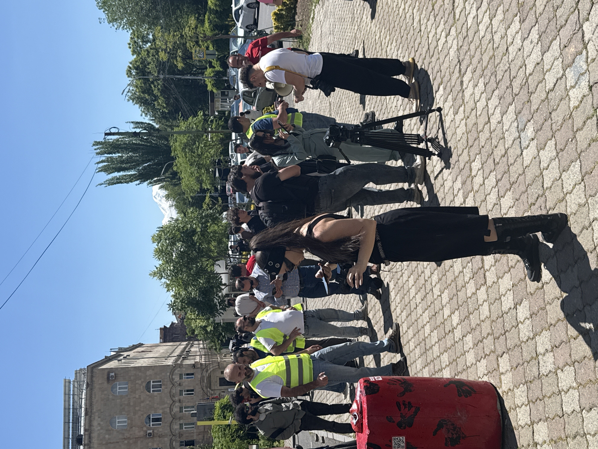 The image captures an outdoor scene on a paved surface, likely a sidewalk or plaza, in Yerevan, Armenia. A group of people are gathered, possibly for a demonstration or protest. There are at least 15-20 individuals visible, some wearing yellow vests. A red barrel is positioned in the foreground with black handprints and a QR code on its side. Buildings are visible in the background, including one with distinctive arched windows. The sky is clear blue. In the distance, a mountain peak is visible above the trees, potentially Mount Ararat. Some people are using cameras and microphones suggesting a media presence. The overall setting is urban, with trees providing some greenery.