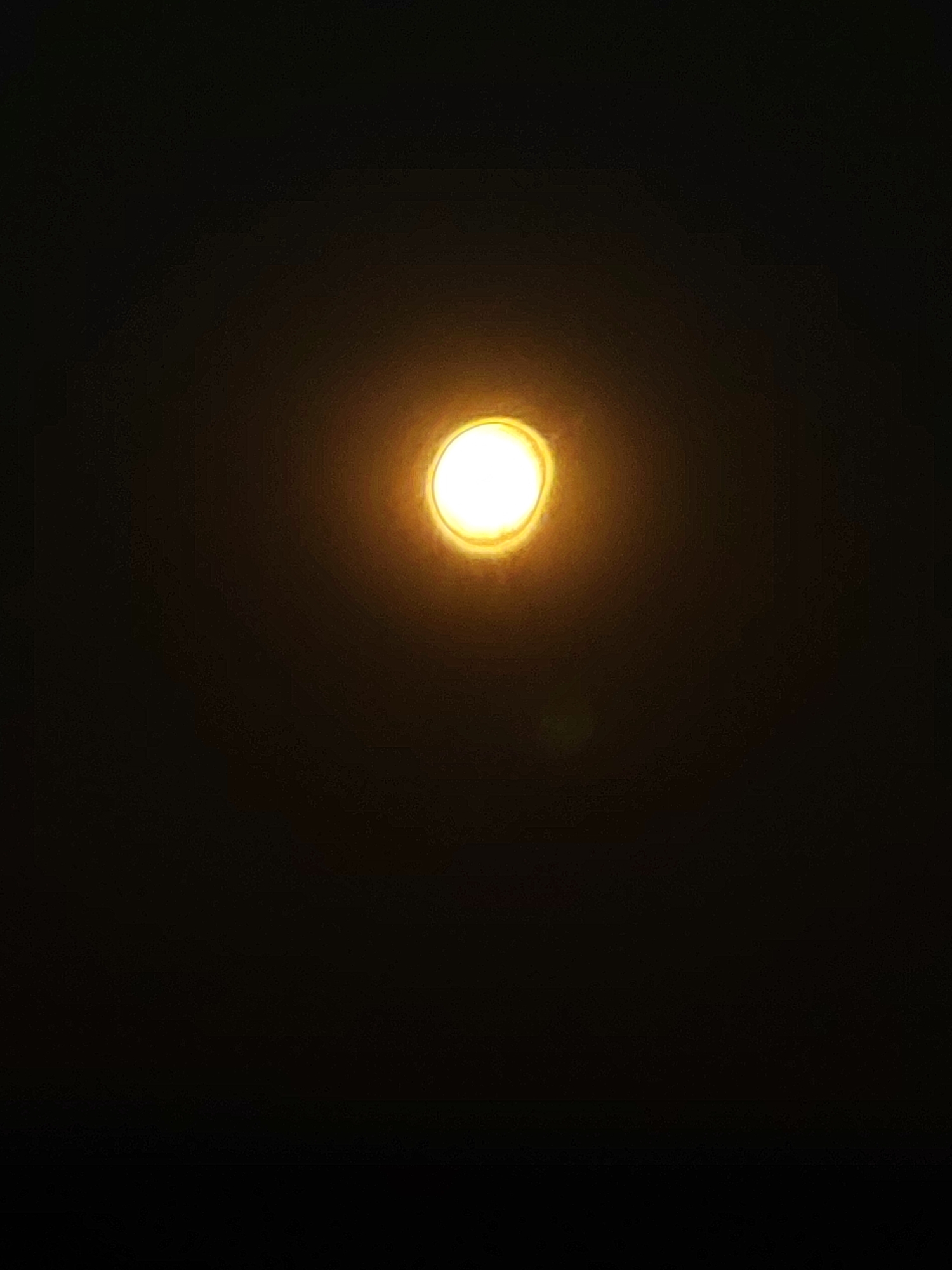 The image is a close-up shot of a single, bright, circular light source against a dark background. The light is predominantly white in the center, with a halo of warm yellow and orange radiating outwards, creating a blurred effect. The overall impression is of a powerful, isolated light. The image does not contain any elements that would allow for identification of a specific location within Autillo de Campos, Spain, or any people, objects, actions, interactions, or environmental details beyond the light source itself.