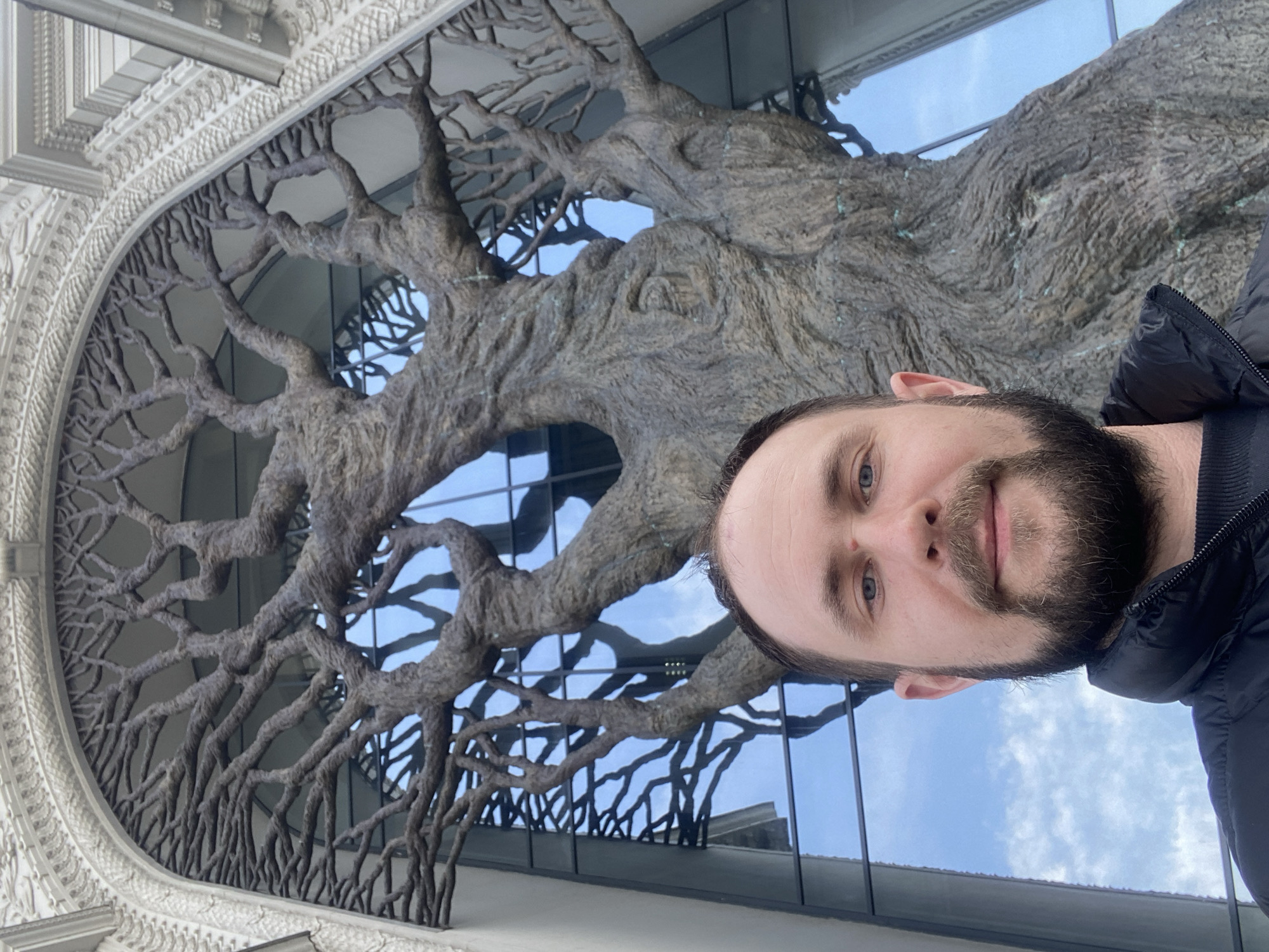 A man with a beard is standing in front of a building with a large tree sculpture integrated into the facade. The building appears to have a modern design with reflective glass panels. The tree is made of a textured, brown material and features numerous branches. The facade has intricate architectural details around the tree sculpture. The man is wearing a black jacket. The sky is visible in the background through the branches of the tree. The location is Kazan, Russia.