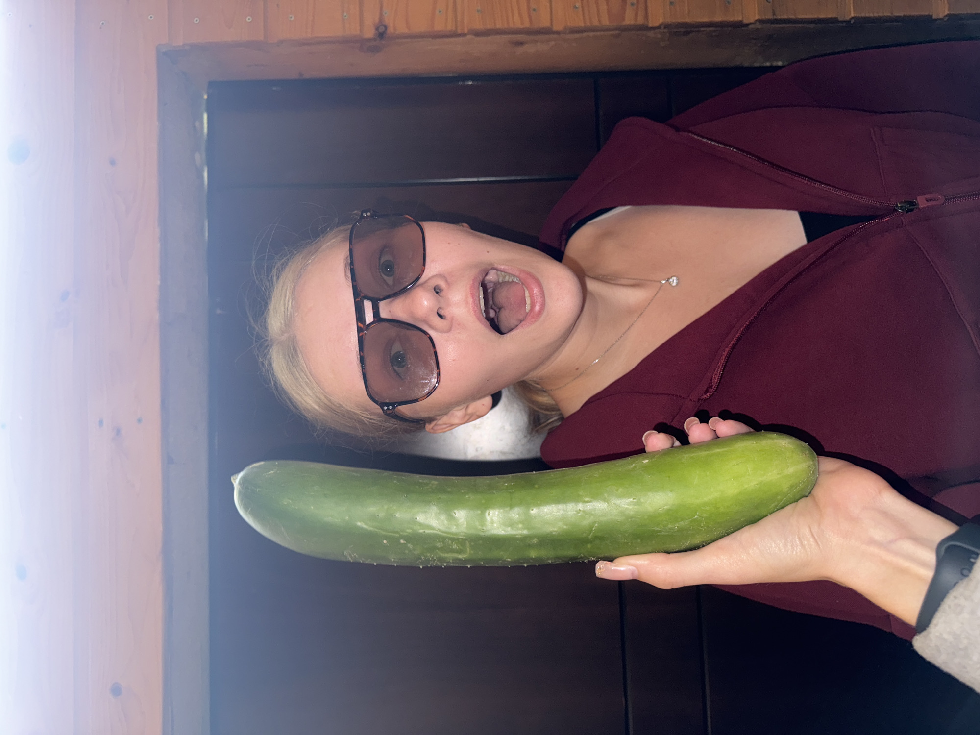 A young Caucasian woman with blonde hair and open mouth is holding a large cucumber. She is wearing large, square sunglasses and a dark red hooded jacket. A silver necklace with a diamond pendant is visible around her neck. The background appears to be a wooden interior, possibly a cabin or a rural dwelling in the village of Borodulino, Russia. The lighting suggests it is indoors.