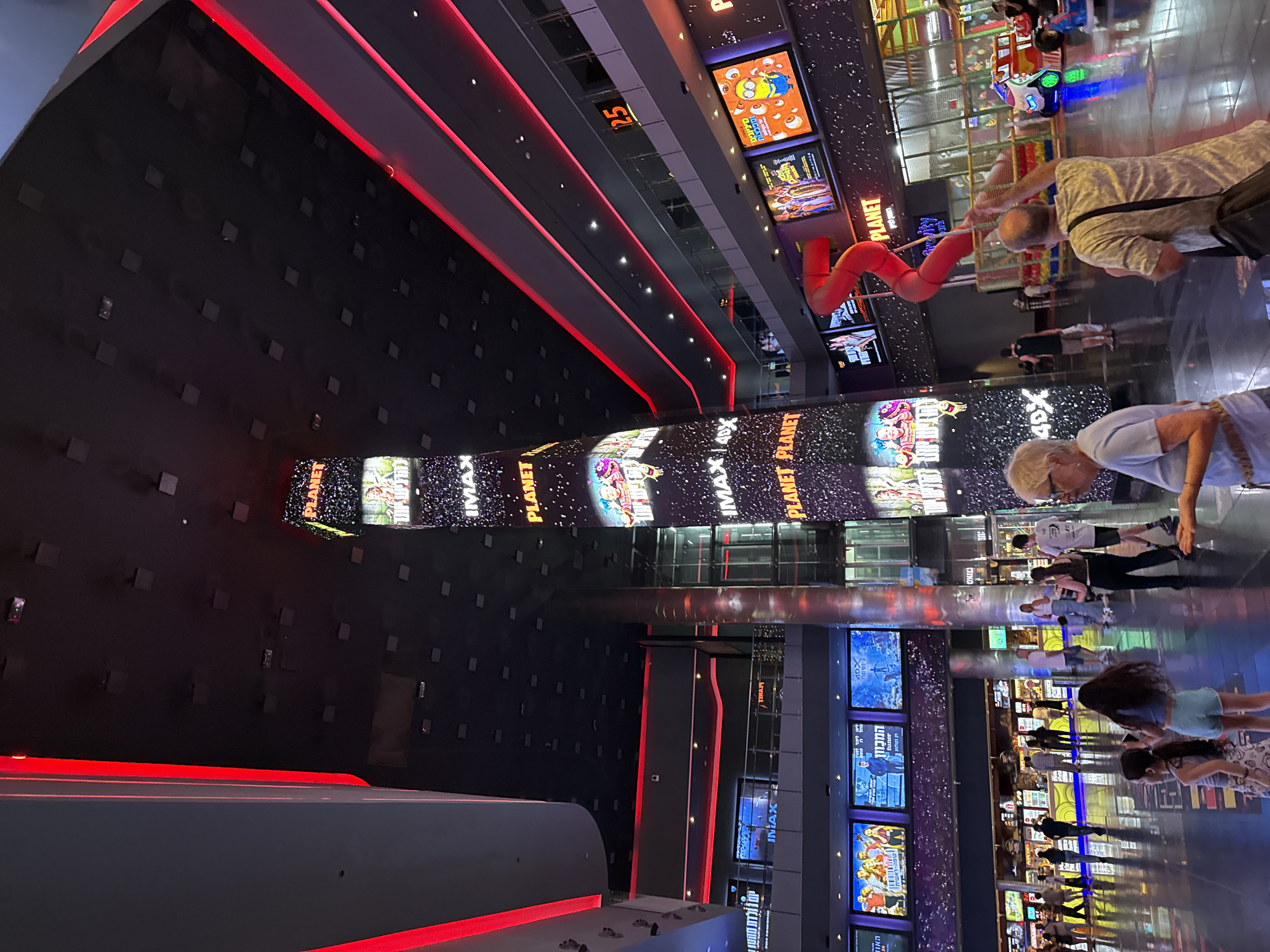 The image depicts the interior of the Planet cinema complex in Rishon LeZion, Israel. The scene is characterized by its modern architecture and vibrant, colorful lighting. A tall, illuminated pillar in the center displays movie advertisements and the "PLANET IMAX 4DX" branding. Red LED lighting streaks along the ceiling and architectural elements, creating a futuristic ambiance.
Multiple large screens are visible throughout the complex, showcasing movie posters and trailers. In the foreground, several individuals are walking and interacting. Two young girls, one in a patterned dress and the other in light blue shorts, are standing. An elderly woman in a light blue shirt and an older man with glasses are also present. In the midground, a concession area with various food and drink offerings is visible, with people queuing. Further in the background, a children's play area with a red slide and rideable toys can be seen. The floor is highly reflective, mirroring the lights and activity. The overall impression is that of a busy and entertainment-focused venue.