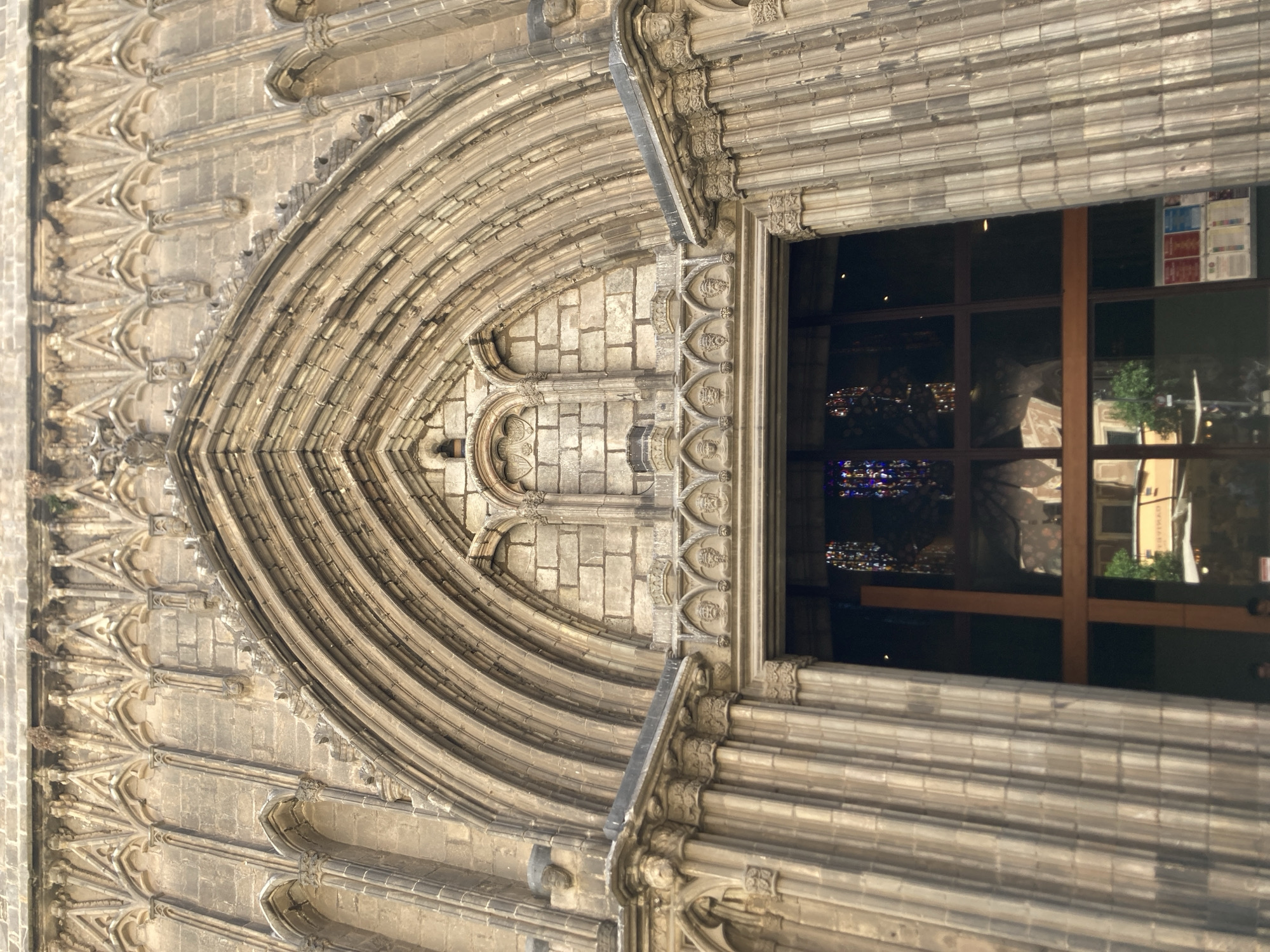 The image shows a low-angle, close-up view of the ornate facade of a gothic cathedral in Barcelona, Spain. The focus is on the grand entrance archway, characterized by intricate stone carvings, multiple layers of ribbed vaults, and decorative elements like small pointed arches and sculpted faces. The stone appears weathered, suggesting significant age. Through the open doorway, stained glass windows with colorful patterns are visible, hinting at the interior's grandeur. Reflections in the glass doors show glimpses of the surrounding city, including buildings and trees. The overall impression is one of imposing historical architecture.
