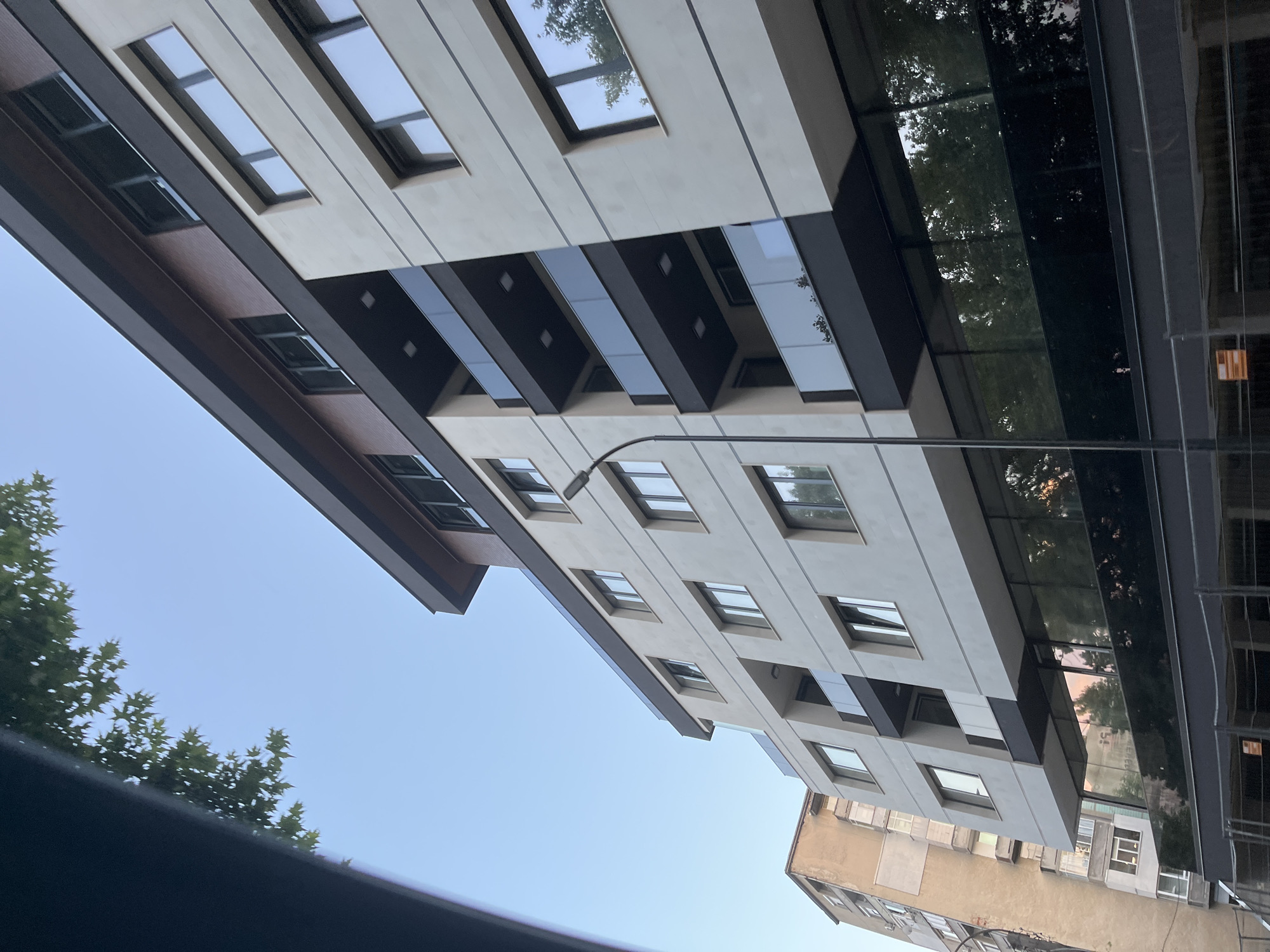 This is a low-angle, vertical-format shot of modern architecture against a clear blue sky. The main subject is a contemporary building with a light-colored facade and dark, horizontal accent strips. The building features numerous windows, some appearing as skylights and others as traditional windows. A striking element is the use of reflective dark glass sections that mirror the surrounding trees and sky, adding depth and a sense of being outdoors.
Adjacent to this modern structure, an older, weathered building with a yellowish facade is visible. This older building has a more traditional appearance with visible brickwork and smaller, more numerous windows. The contrast between the two buildings highlights different architectural eras and styles.
Green trees with dense foliage are visible, with some branches extending into the frame, their leaves reflecting in the glass surfaces of the modern building. A street lamp extends from the modern building into the frame, suggesting an urban or semi-urban setting. The sky is a consistent, clear blue, indicating fair weather. The lighting suggests it is daytime, possibly late afternoon given the quality of light reflected in the glass. There is no visible text in the image. The overall mood is one of urban development and the juxtaposition of old and new.