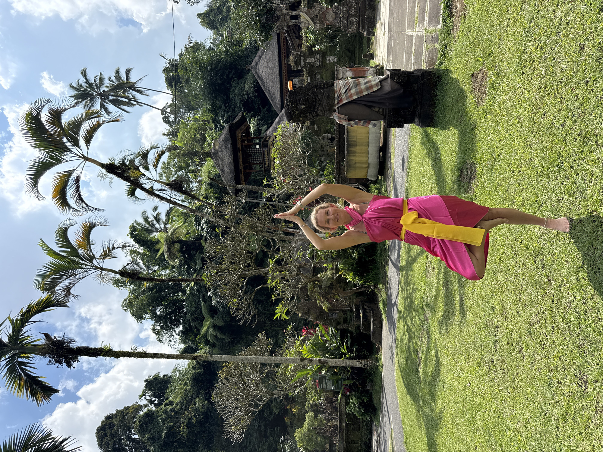 In Tegallalang, Indonesia, a woman stands on a verdant lawn in a tree pose, a yoga posture. She is dressed in a vibrant pink wrap dress adorned with a yellow sash. Her hands are pressed together above her head, palms touching, and her left leg is bent, balancing on the ball of her foot, while her right leg is extended behind her. The woman's blond hair is pulled back, and she gazes forward with a serene expression.
Behind her, a paved path curves through the well-maintained garden. Lush green grass covers the ground, dotted with the shadows of palm trees that tower overhead. The background is a dense jungle canopy, with a glimpse of traditional Balinese architecture featuring thatched roofs and stone carvings. The sky above is a brilliant blue, punctuated by fluffy white clouds. The scene evokes a sense of tranquility and natural beauty.