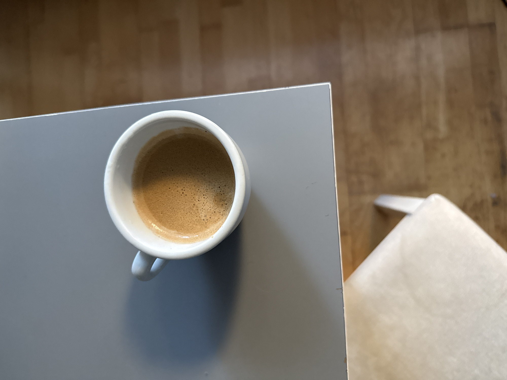 A white ceramic espresso cup with a light blue handle is positioned on the left side of a flat, gray surface. The cup is filled with dark brown espresso, topped with a layer of crema that shows small bubbles. A shadow of the cup is cast to the left. The gray surface is bordered by a wooden floor with visible grain patterns on the right side. In the foreground, a portion of a light-colored chair is visible, angled towards the right. The image does not provide any information to indicate the location is the city of Wrocław, Poland.