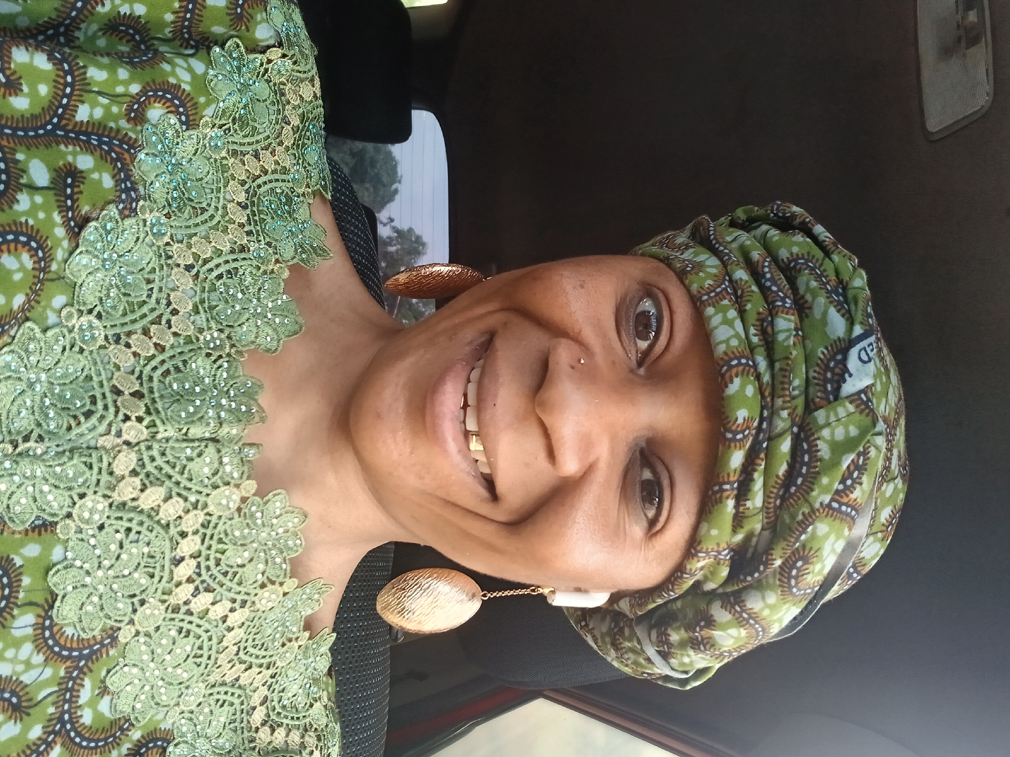 A smiling, dark-skinned woman is pictured from the chest up, inside a car. She wears a patterned green and brown headscarf and a green dress with lace trim. Large, gold-colored earrings hang from her ears. She appears to have a nose piercing. The car's interior is visible behind her, including the dashboard and window. The location is Rigachikun, Nigeria.
