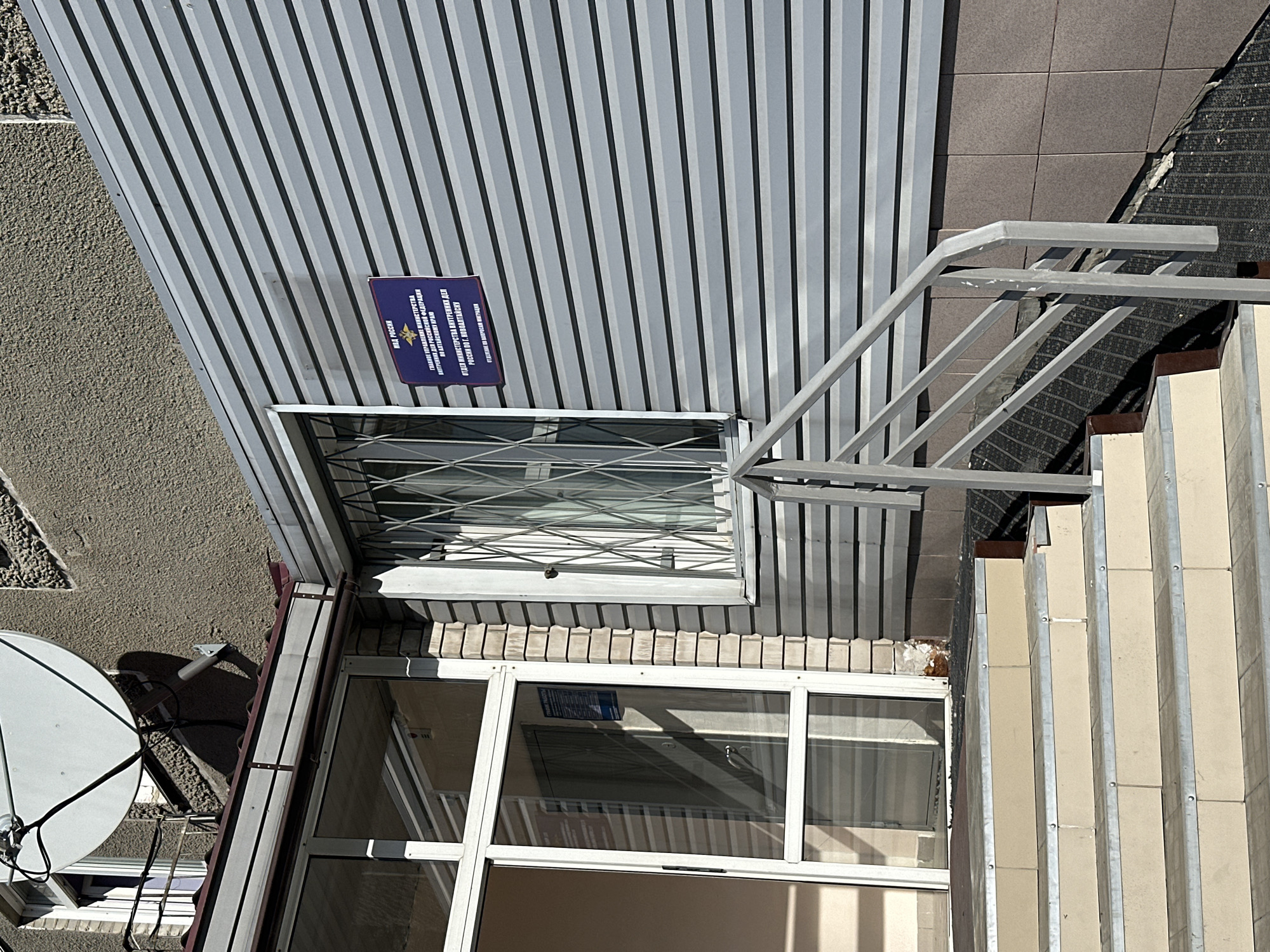 The image shows the exterior of a building in Novoaltaysk, Russia, featuring a light-colored tiled staircase leading up to an entrance with glass doors and windows. To the right of the entrance, a silver-colored corrugated metal siding covers the wall, with a blue sign indicating "МВД РОССИИ" (Ministry of Internal Affairs of Russia) and further text in Russian, including "ГЛАВНОЕ УПРАВЛЕНИЕ МИНИСТЕРСТВА ВНУТРЕННИХ ДЕЛ РОССИЙСКОЙ ФЕДЕРАЦИИ ПО АЛТАЙСКОМУ КРАЮ" (Main Directorate of the Ministry of Internal Affairs of the Russian Federation for the Altai Krai) and "ОТДЕЛ МИНИСТЕРСТВА ВНУТРЕННИХ ДЕЛ РОССИИ ПО Г. НОВОАЛТАЙСКУ" (Department of the Ministry of Internal Affairs of Russia for the city of Novoaltaysk). A barred window is visible to the right of the sign. Above the corrugated siding, a textured concrete wall is visible, with a satellite dish mounted on the upper section. A metal handrail runs alongside the staircase and a ramp. The environment appears to be a public building in a city setting.