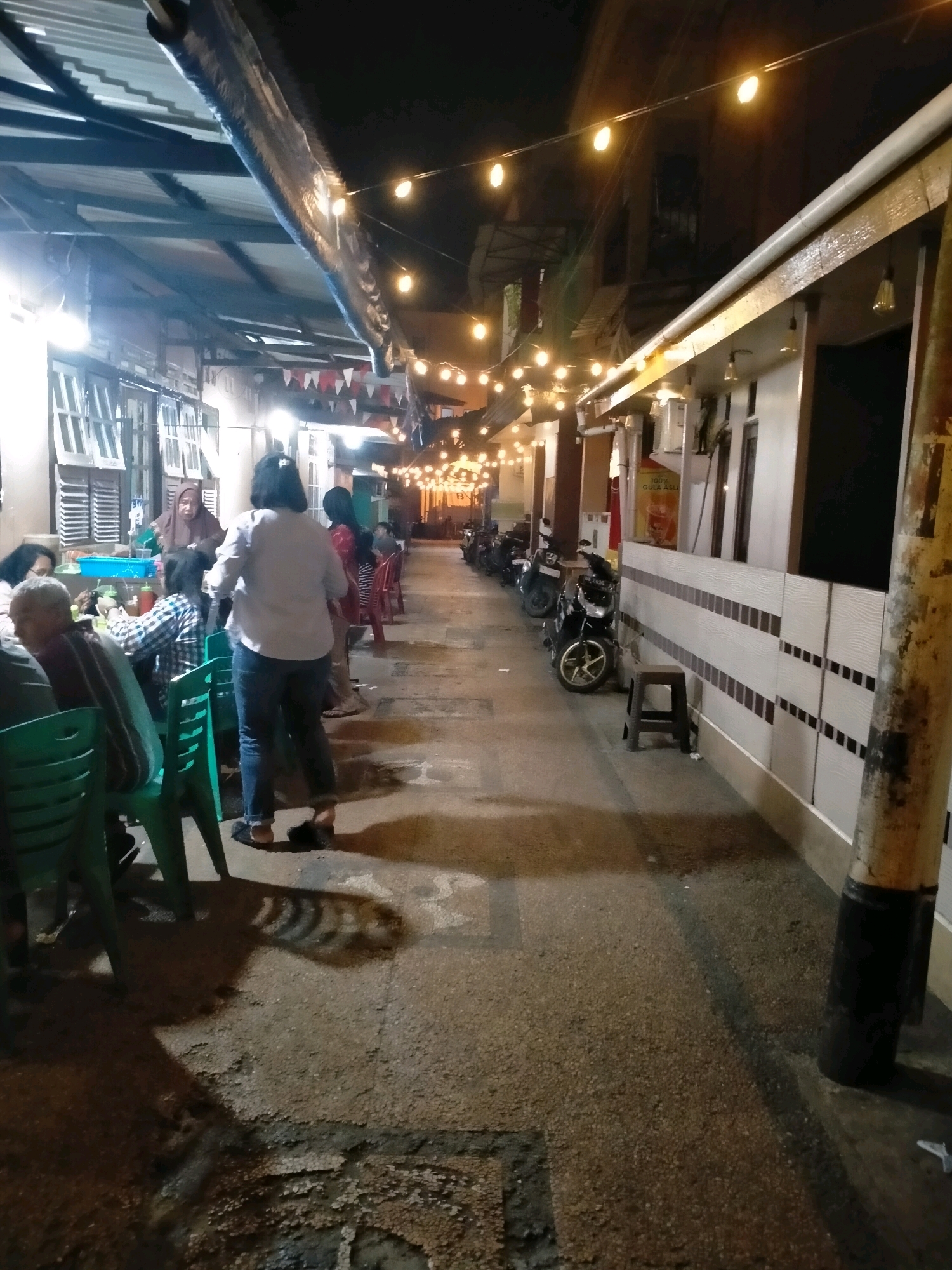 A group of people are seated at tables under a canopy illuminated by string lights, located on a street in Ambon, Indonesia. The scene depicts an outdoor dining area, likely a food stall or small restaurant, with several motorcycles parked along the side of the street. A woman in a headscarf and a woman in a white shirt and jeans are standing and interacting near the tables. Green plastic chairs are arranged around the tables. The street surface is paved with asphalt and shows some wear and tear. The buildings in the background suggest a densely populated urban environment. The overall atmosphere is that of a casual evening gathering for food and socializing.