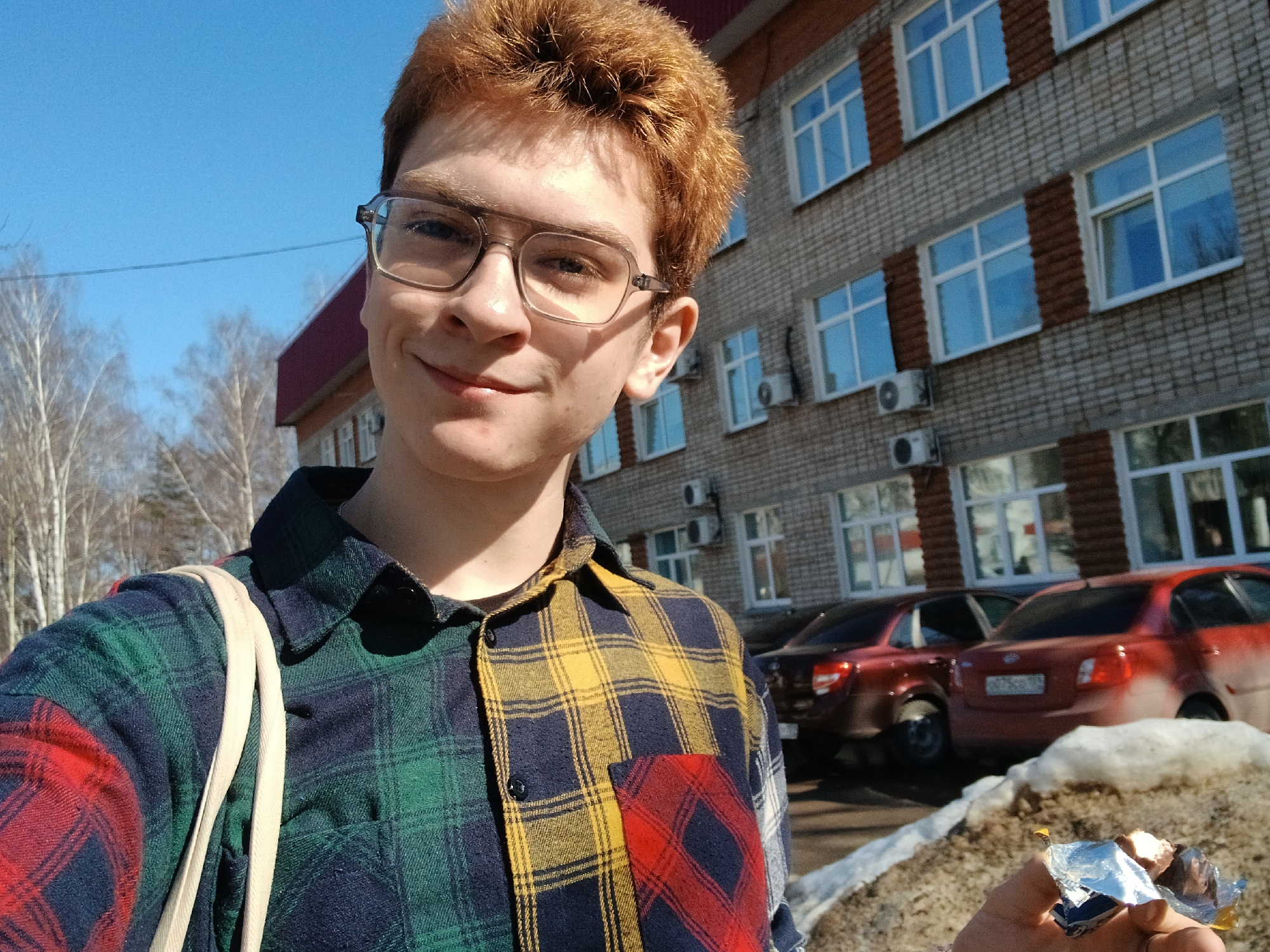 A young person with reddish-brown hair and glasses is taking a selfie outdoors in Chaikovsky, Russia. They are wearing a colorful patchwork plaid shirt. Behind them is a multi-story brick building with multiple windows and air conditioning units. In the background, there are several parked cars, and the ground has patches of snow and dirt. The sky is blue and sunny. In the person's hand, there appears to be wrapped candy.