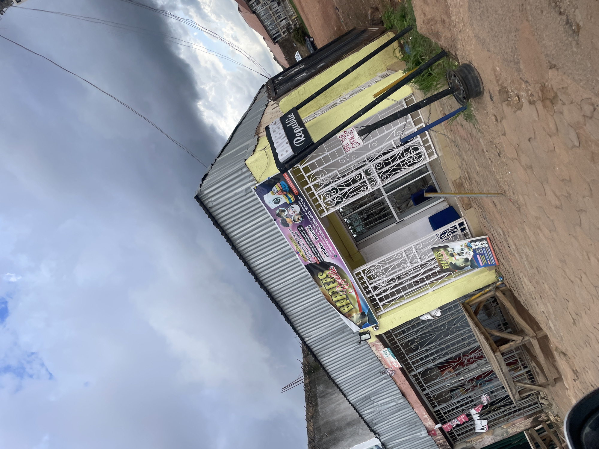 The image shows a street view of a barbershop called "Republic Barbers" in Gura Top, Nigeria. The shop is painted yellow and has a corrugated metal roof. There are various signs advertising the services offered, including haircuts for men, women, and children, as well as pedicures, laundry services, and phone accessories. The shop appears to be open, with decorative metal gates covering the windows.
The time of day is difficult to ascertain, but the lighting suggests it could be daytime. The sky is cloudy, indicating overcast weather. The street in front of the shop is paved with uneven stones and dirt. There is also a discarded tire propped up on a metal stand outside the shop. The overall impression is a bustling local business in a developing area.
Visible text includes service offerings like "HAIRING, PEDICURE COSMETICS, LAUNDRY SERVICES & PHONE ACCESSORIES," operating hours "8:00AM 9:00PM," and contact phone numbers. There's also text related to specific services like "CHILDREN CUTS, ADULTS CUTS LADIES CUTS, RELAXER & DYE," and "DREAD LOCKS, LAUNDRY SERVICES, PEDICURE & LOTS MORE." The location is further specified as "SHOP NO 12 ZARAMANG CADA RAYFIELD ROAD ENTRANCE TO KALWA HOUSE."
