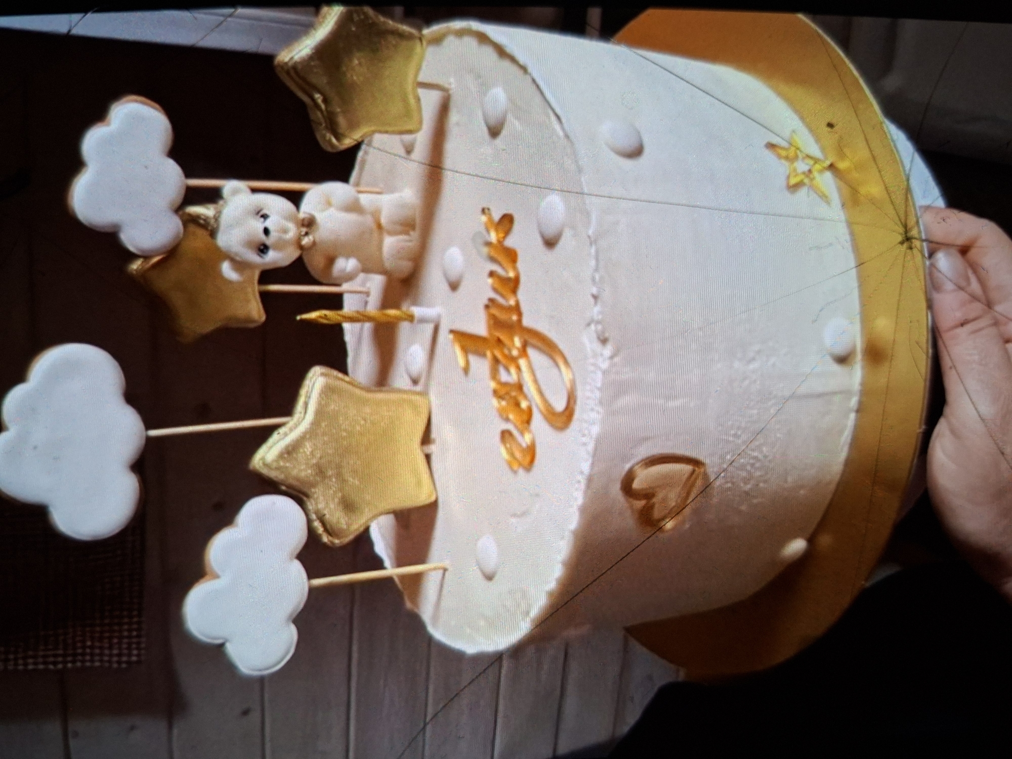 This image shows a white round cake decorated with gold accents, a fondant teddy bear, and gold stars. White cloud-shaped decorations are also attached to skewers, along with the stars and the teddy bear, giving the impression of a sky theme. The cake has gold lettering on its side, which appears to spell out a name, possibly "Luna" or a similar word. The cake sits on a gold cake board. A person's hand is visible on the right side of the frame, holding the edge of the cake board. The background is blurred but appears to be indoors, with a wooden texture suggesting a table or shelf. The lighting is artificial and appears to be coming from above. The overall impression is a celebratory cake, likely for a birthday or baby shower, given the whimsical decorations. No specific time of day or weather conditions are discernible.