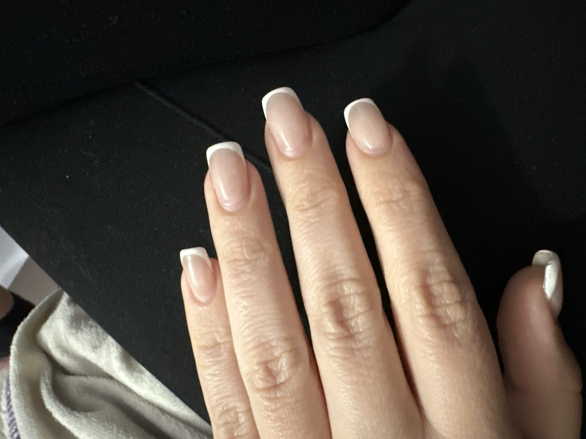 This close-up, eye-level shot features a human hand, specifically a woman's left hand, presented against a soft, dark fabric background, with a lighter, textured fabric visible in the lower left corner. The hand is the central subject, meticulously cared for with a classic French manicure. Each nail is long, with a distinct square or squoval shape, painted with a natural, translucent pinkish base and crisp white tips. The skin on the fingers and palm shows natural creases and texture. The fingers are slightly spread, showcasing all five nails, though the thumb nail is partially obscured. The lighting is soft and even, highlighting the glossy finish of the nails. No activity is depicted beyond the presentation of the hand and its manicure. There are no discernible time of day or weather cues, and no visible text. The setting appears to be an indoor, personal environment. This image was captured in Sofia, Bulgaria.