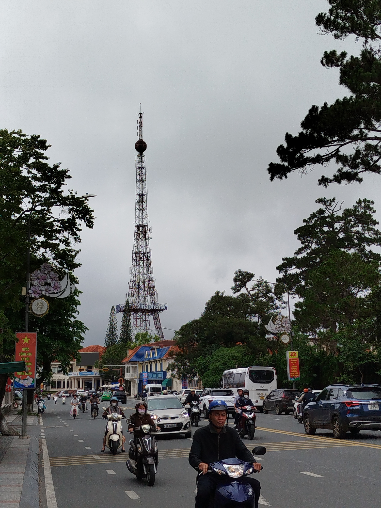 The image depicts a busy street scene in Da Lat, Vietnam, under an overcast, daytime sky. A tall, lattice-structured communication tower, topped with a spherical element and displaying the "Vinaphone" logo, is the dominant feature in the background, towering over the city's architecture.
The street is bustling with traffic, primarily motorcycles and scooters, with several cars interspersed. Riders on most motorcycles wear helmets, and some also wear face masks. A dark blue SUV is visible on the right, alongside other vehicles.
Along the left side of the road, a sidewalk is lined with green trees and a decorative street light adorned with floral motifs. A red banner, featuring a yellow star and Vietnamese text, hangs from a pole, signifying a national presence. Buildings lining the street display European-influenced architecture, common in Da Lat, characterized by pitched roofs and detailed facades. Lush green foliage, including tall pine trees, frames the upper and side portions of the image.
The visible text includes:
- On the red banner: "NƯỚC CỘNG HÒA XÃ HỘI CHIA MUÔN NĂM!"
- On the communication tower: "Vinaphone"
- On vehicle license plates: "49A-07093", "84E-004.92", and "48A 195.57".
The scene captures the daily activity and urban landscape of Da Lat.