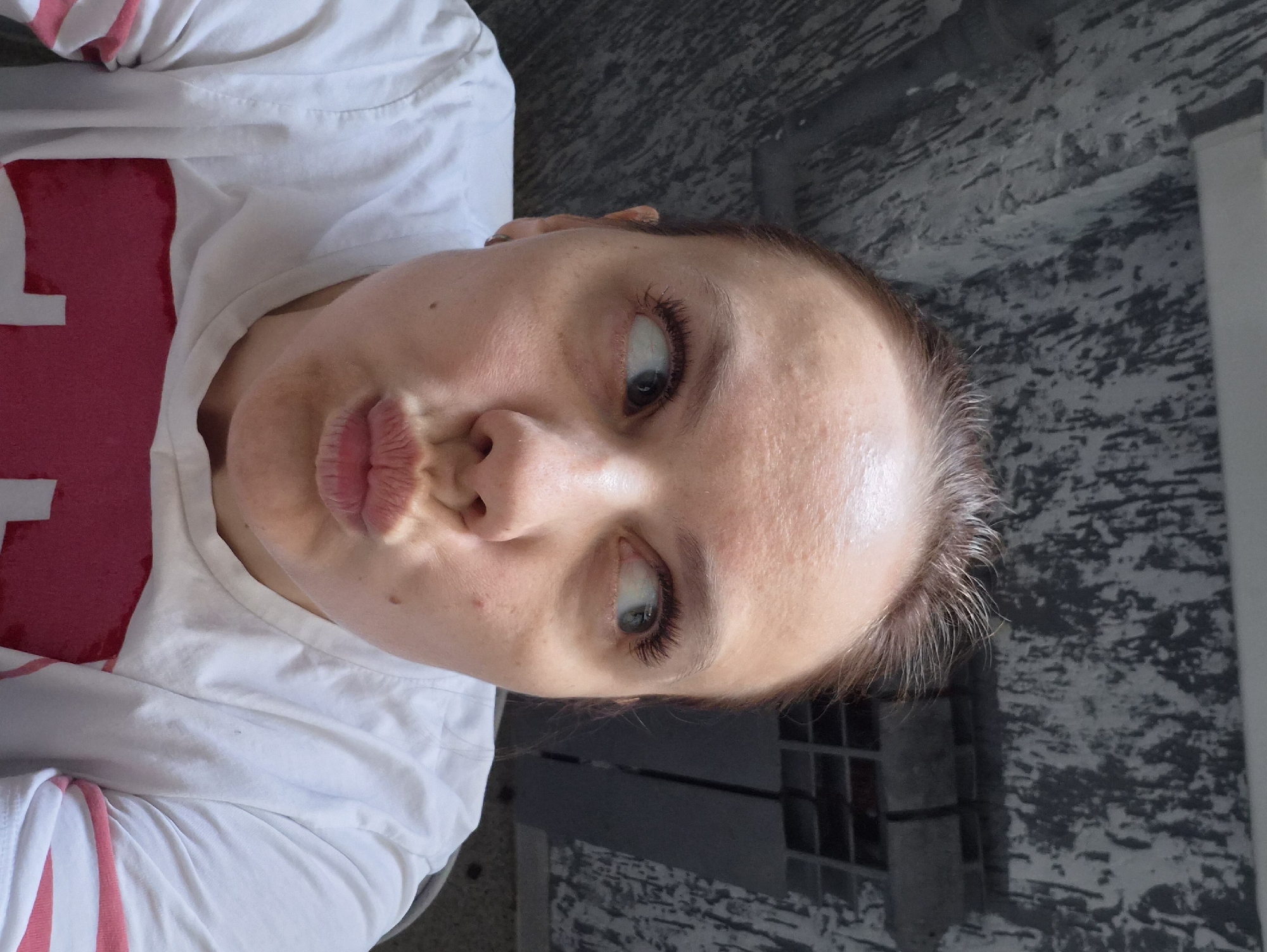 A Caucasian female with light skin and light brown hair is positioned at an angle in the image, wearing a white t-shirt with pink lettering. She is making a "kissing" face. Behind her, a textured wall of grey, black, and white can be seen. To the right, a dark grey object that looks like a bench and a window with several panes are visible. The image appears to have been taken outdoors in daylight. It is impossible to verify that this image was taken in the city of Chaikovsky, Russia.