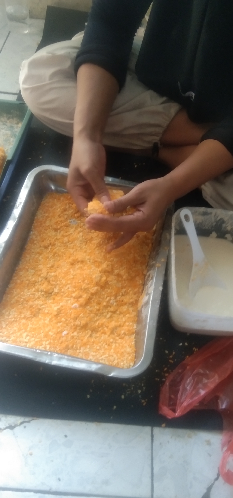 The image depicts a close-up, overhead view of a person's hands preparing food, likely in Rahayu, Indonesia. The individual is seated, wearing light-colored pants and a dark long-sleeved shirt. Their hands are engaged in coating small pieces of food, which appear to be a light-colored dough or batter, with a generous amount of bright orange breadcrumbs or a similar coating. These pieces are being added to a rectangular metal tray filled with the same orange coating. To the right of the tray of breadcrumbs, there is a white plastic container holding a pale, creamy liquid and a white plastic spoon. A crumpled red plastic bag is visible in the lower right corner of the frame. The surface beneath the food preparation area appears to be dark, possibly a countertop, with a tiled floor visible at the bottom. The lighting suggests an indoor setting, possibly a kitchen or food preparation space.