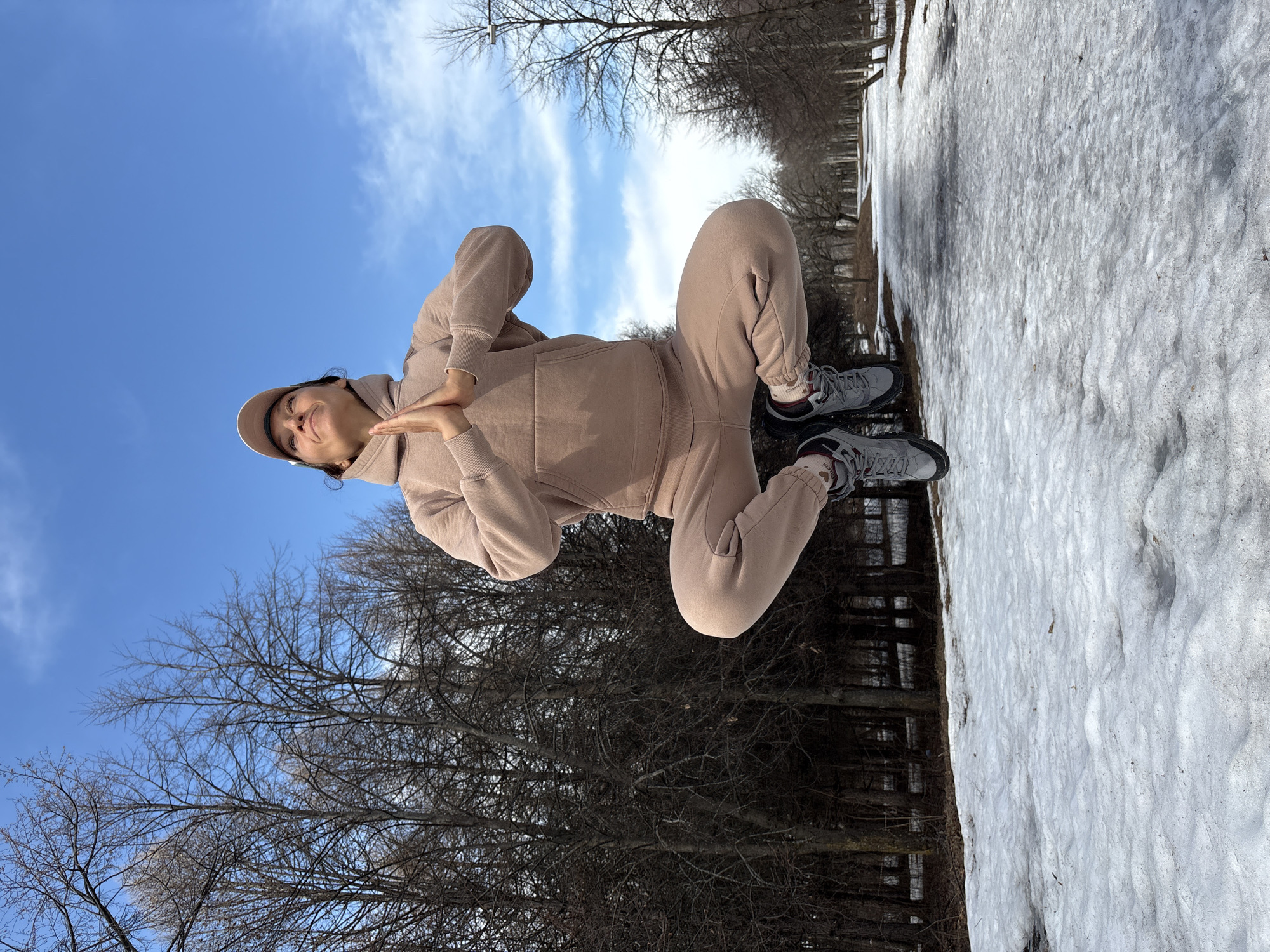 A person wearing a beige tracksuit and cap is performing a yoga pose outdoors. The backdrop includes a blue sky with some clouds, bare trees, and patches of snow on the ground. The location appears to be a park or green space within the city of Ulyanovsk, Russia, given the urban environment mixed with natural elements. The person is standing near a pathway covered in melting snow.