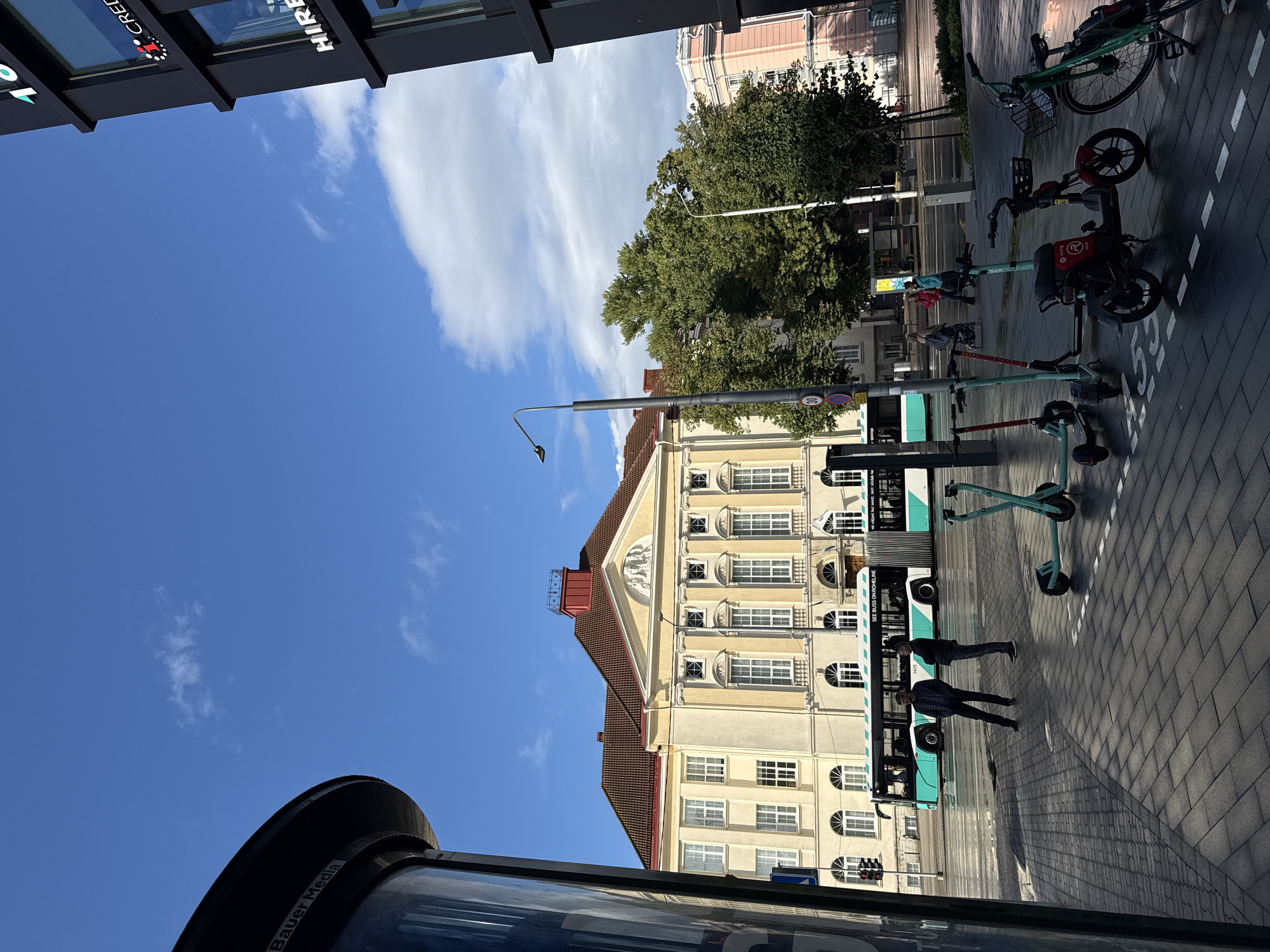 The image shows a street scene in Tallinn, Estonia, on a sunny day with scattered clouds. In the foreground, there is a paved pedestrian area with several parked electric scooters and a bicycle. Two men are standing on the pedestrian walkway, facing each other.
Behind them, a modern articulated bus with a teal and white livery is stopped at a bus stop. The bus has "SEE BUSS ON ROHELINE" printed on its side. Across the street, a grand, light-yellow building with a red-tiled roof dominates the background. This building appears to be a historical or institutional structure, possibly the Town Hall or a university building.
To the right of the scene, a dark, modern building with large windows is visible. Signs on this building include "CREDI" and "HIRE". The street appears to be wet, suggesting recent rain. Trees line the street behind the bus, adding greenery to the urban landscape. The sky is a bright blue with wispy white clouds.